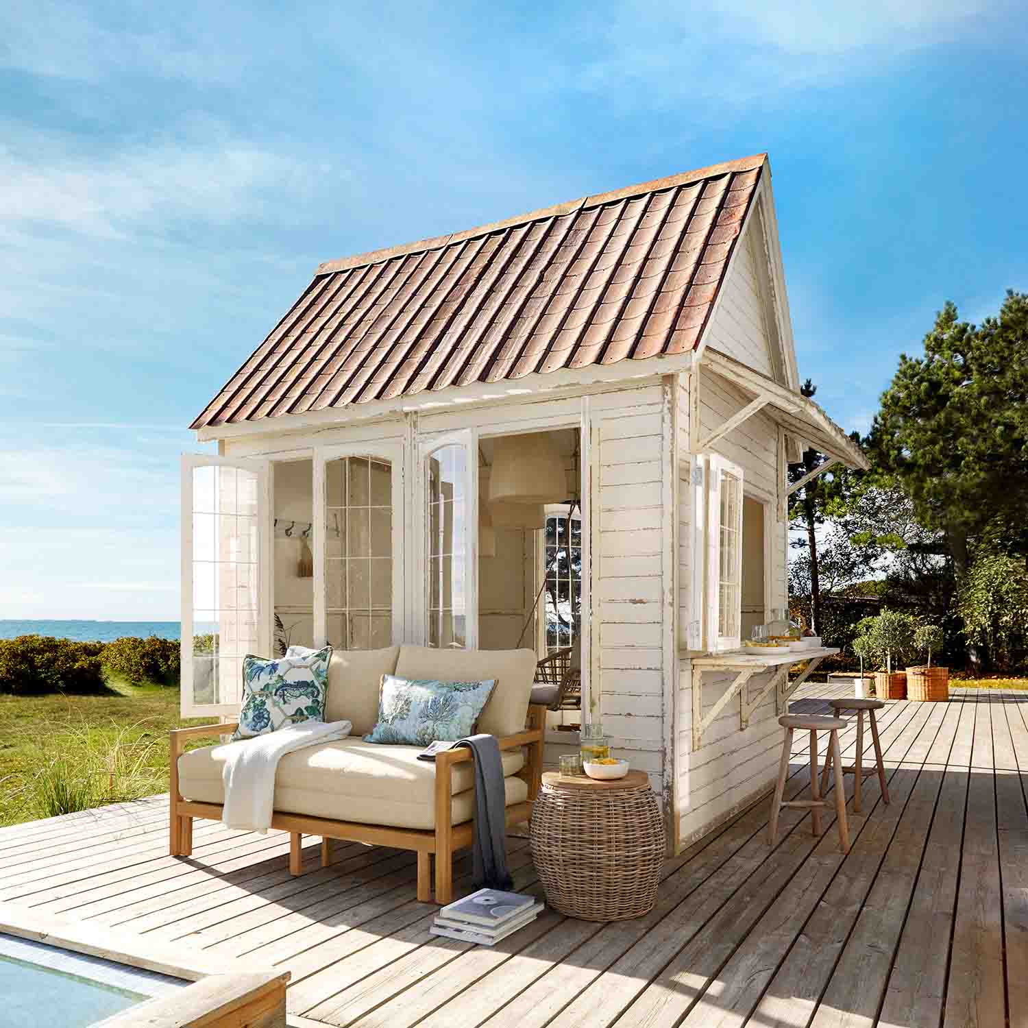 Kiosque en bois blanc avec toit rouge sur terrasse en bois avec canapé beige et table en osier