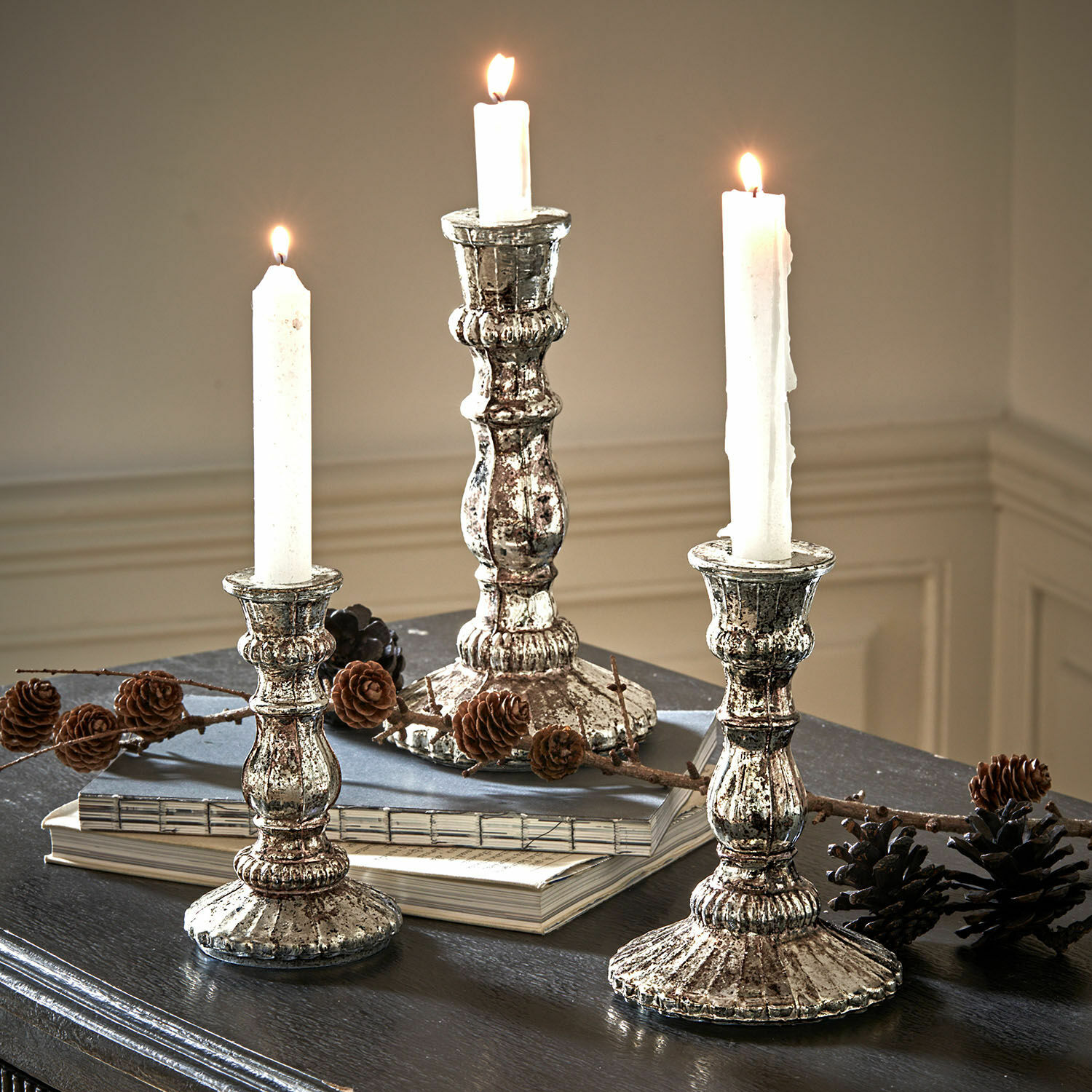 Kandelaar set van 3 Favalina Drie vintage metalen zilveren kandelaars met witte kaarsen op een donkere tafel met dennenappels