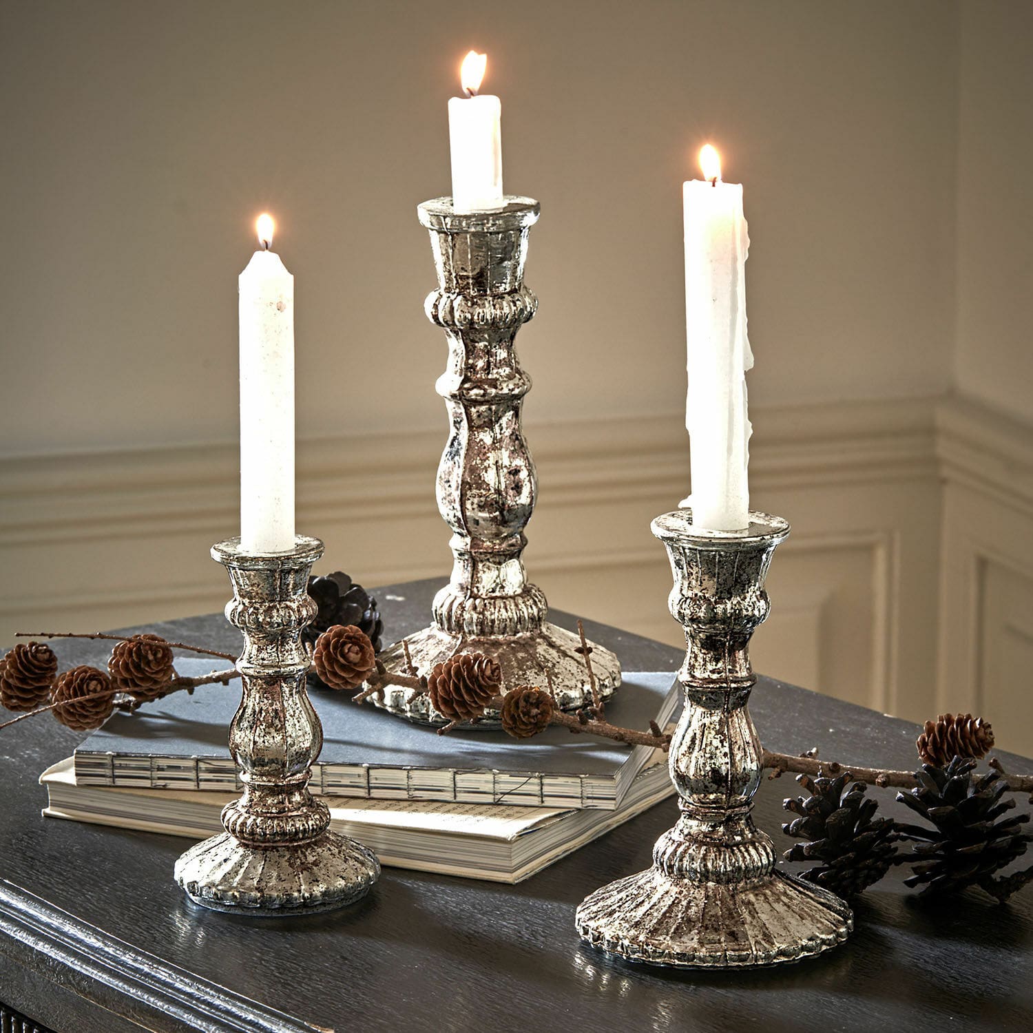 Kandelaar set van 3 Favalina Drie vintage metalen zilveren kandelaars met witte kaarsen op een donkere tafel met dennenappels