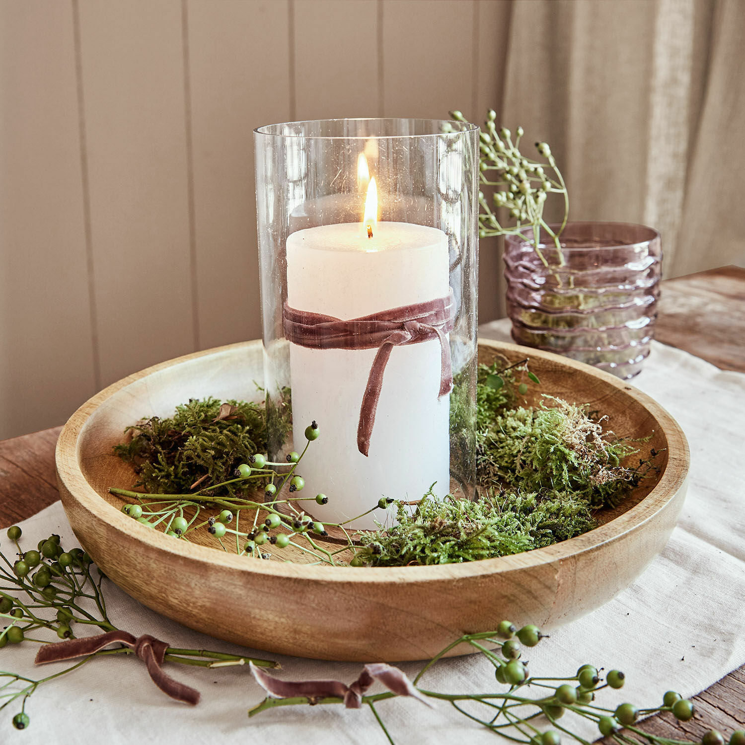 Witte kaars in glazen houder met fluwelen lint op houten schaal met groen mos versierd