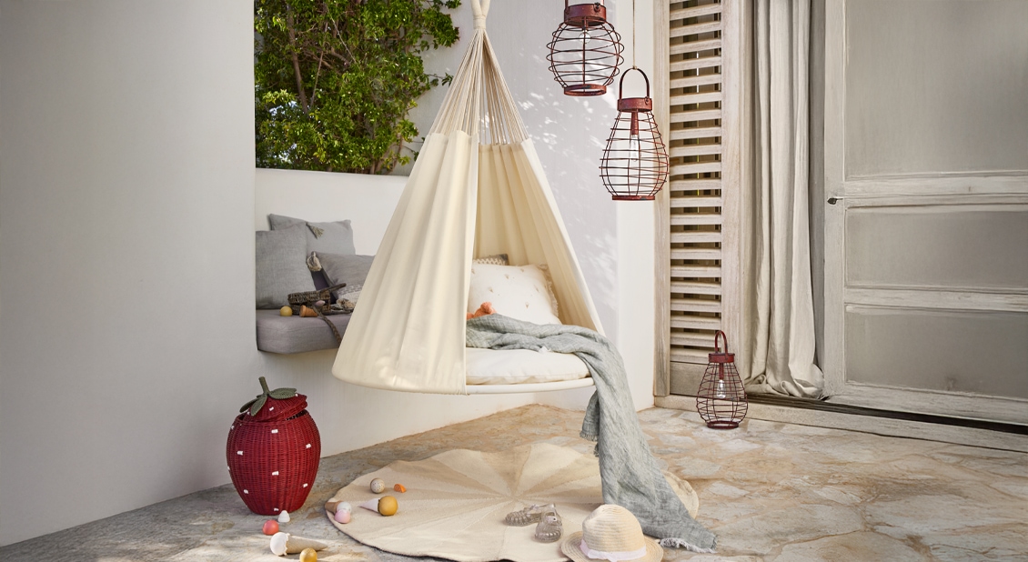 Beige hangende stoffen schommel met kussens en grijze deken op terras, gezellige tuindecoratie