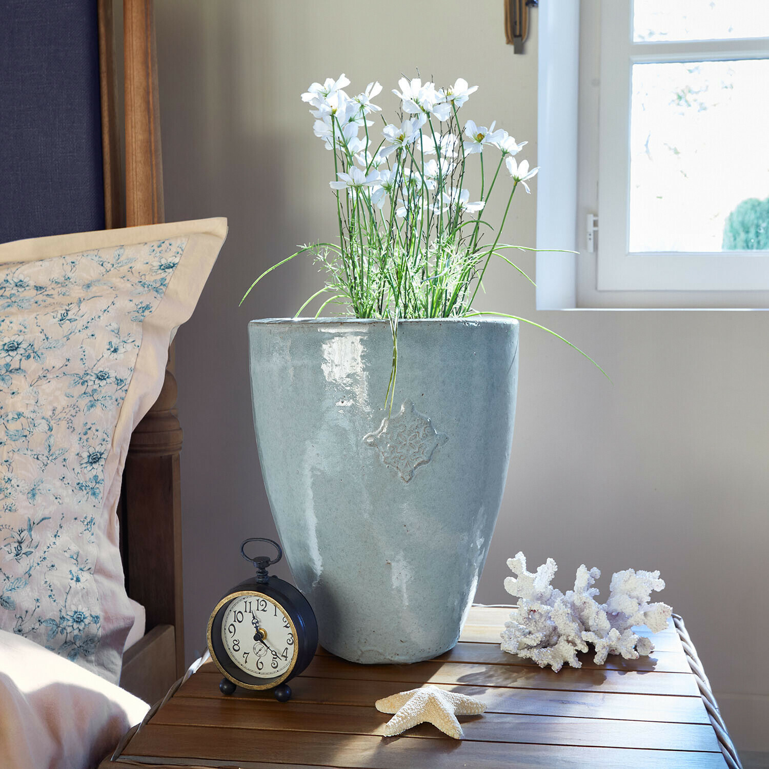 Grijze keramische bloempot met witte bloemen op houten tafel naast wekker en zee decoraties