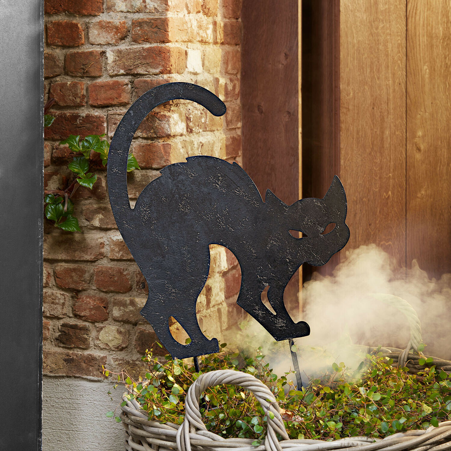 Tuinsteker Rénelise Decoratieve zwarte metalen kattenfiguur in een mand met groene planten voor een bakstenen muur