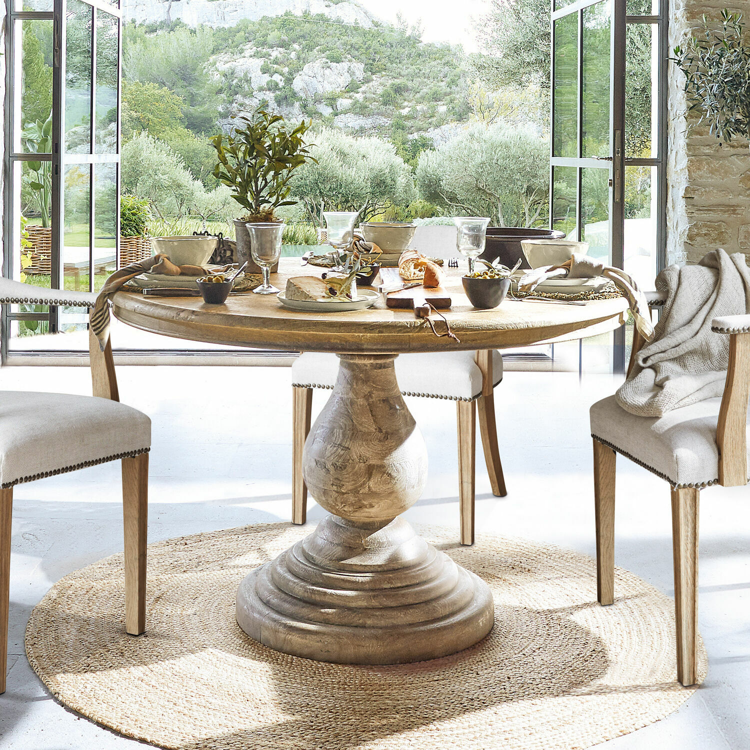 Table Orlin Table ronde en bois avec pied sculpté sur tapis naturel dans une salle à manger lumineuse avec vue sur le jardin