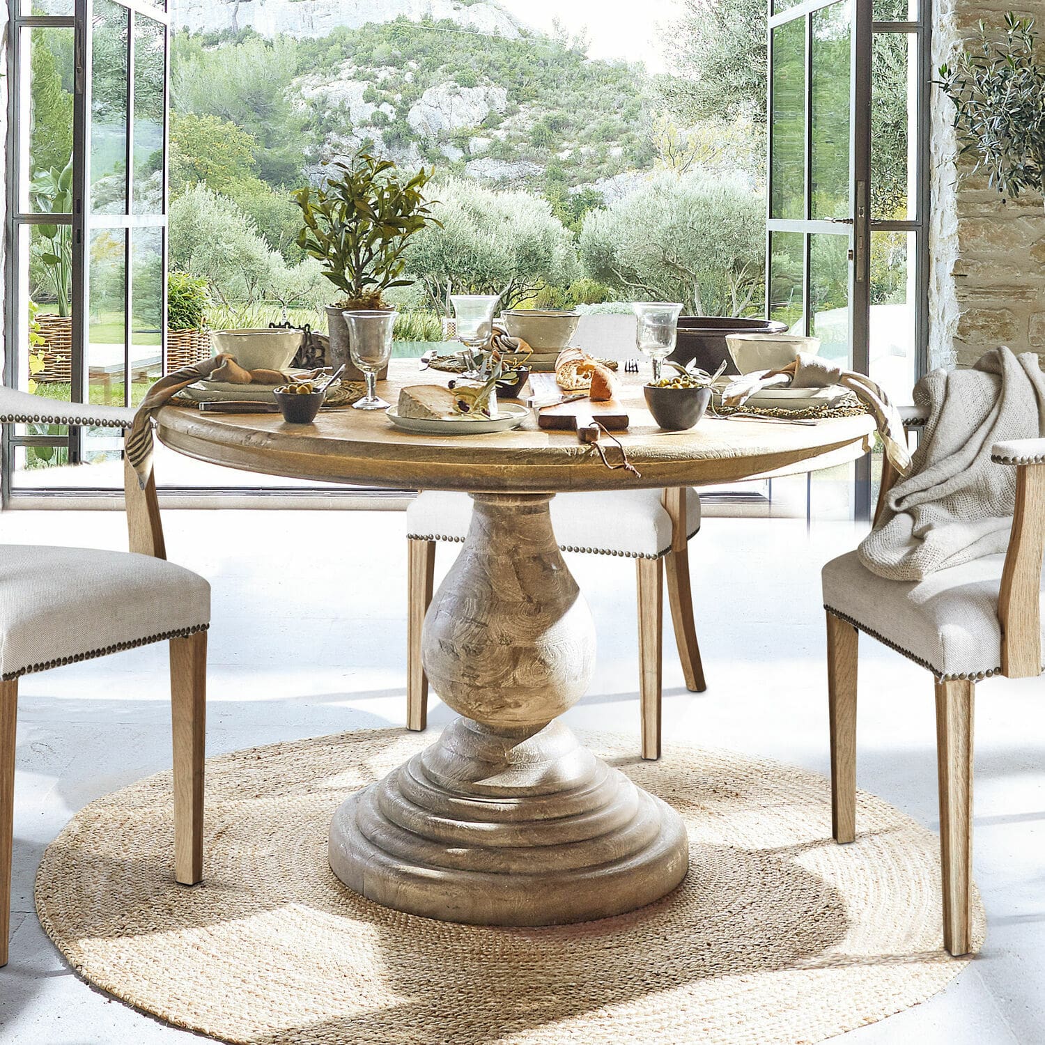Table Orlin Table ronde en bois avec pied sculpté sur tapis naturel dans une salle à manger lumineuse avec vue sur le jardin