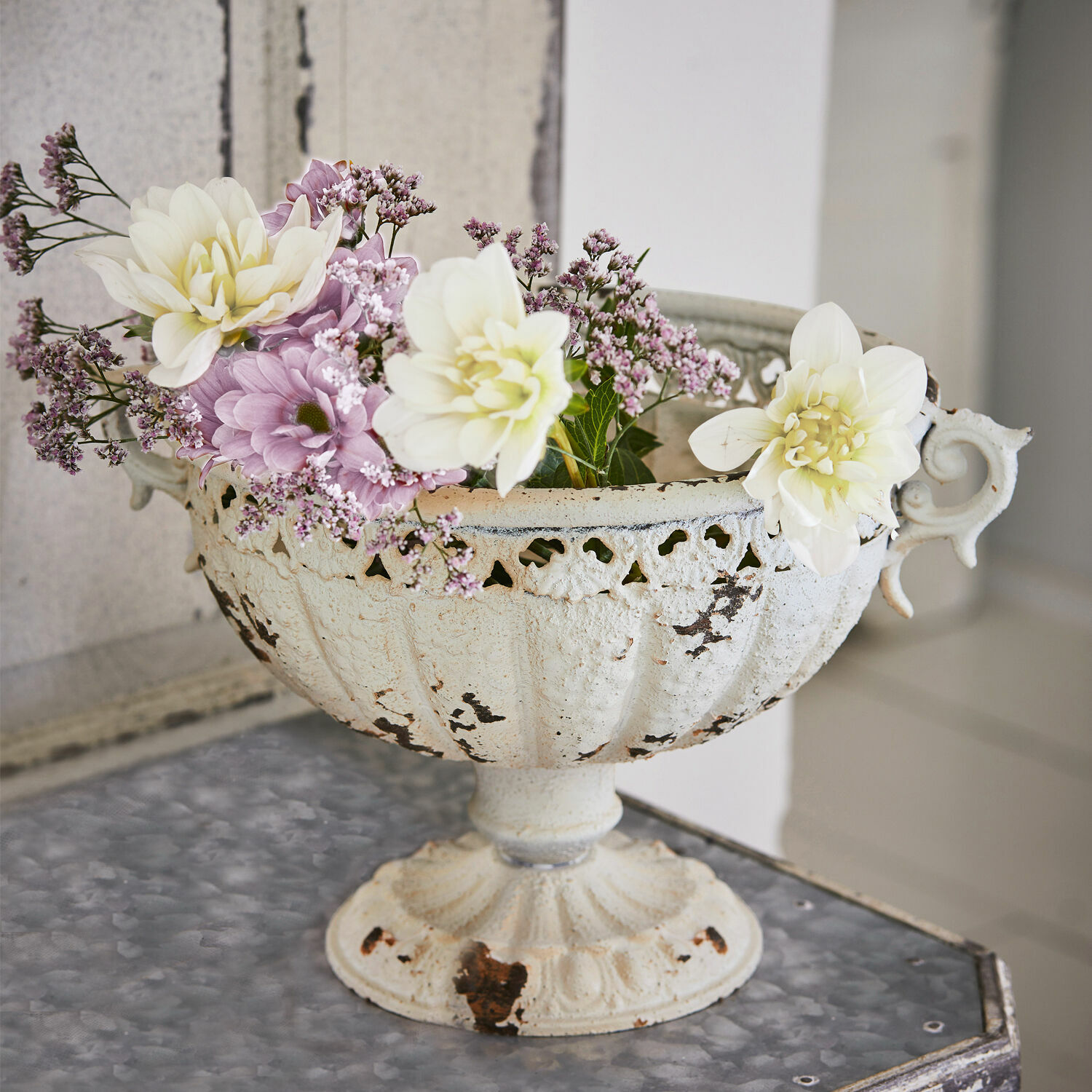 Coupe Vescemont Coupe en métal blanc vieilli avec fleurs pastel posée sur une table décorative intérieure