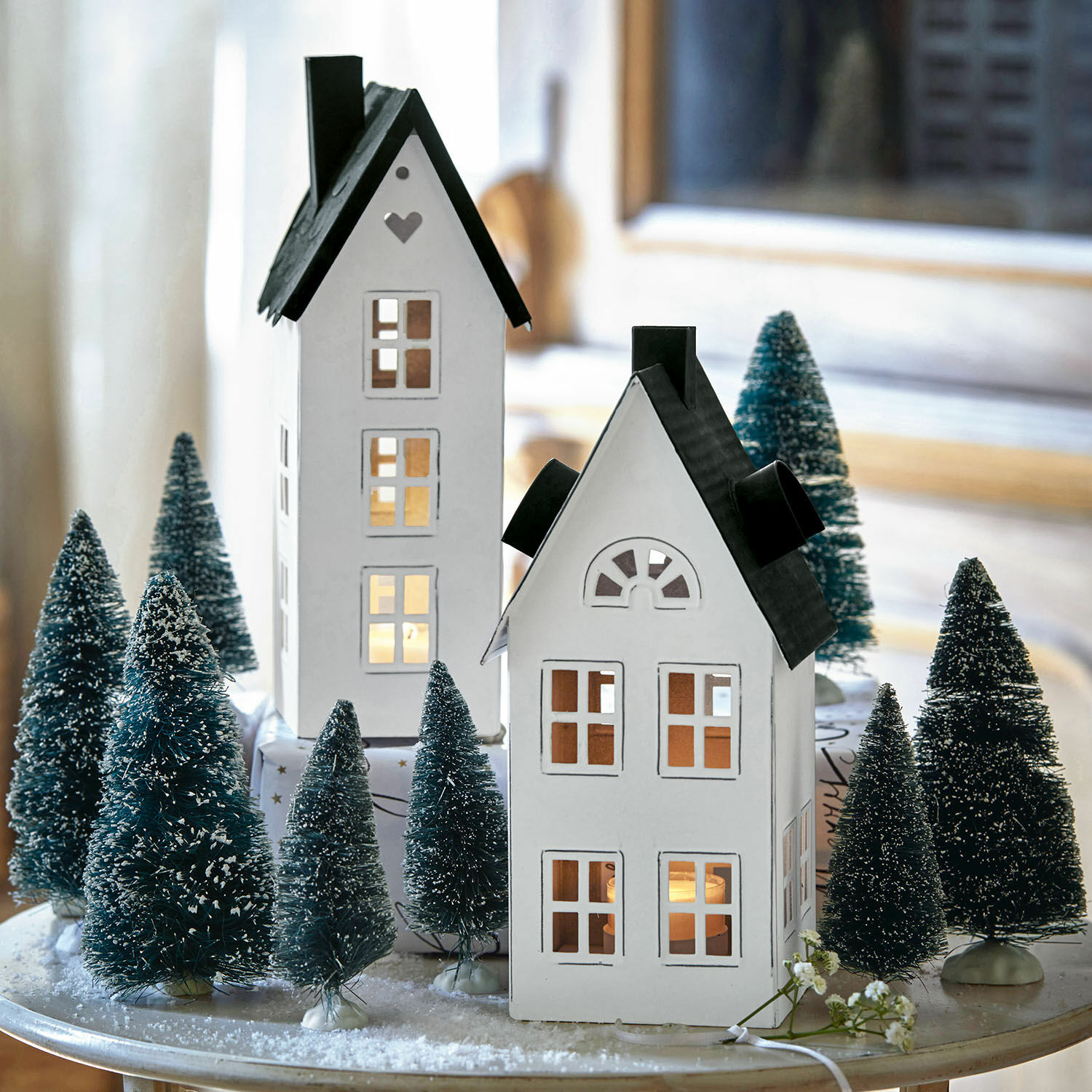 2 lanternes maisonnettes Gunar Maisons décoratives blanches en métal avec toits noirs entourées de sapins artificiels sur une table de Noël