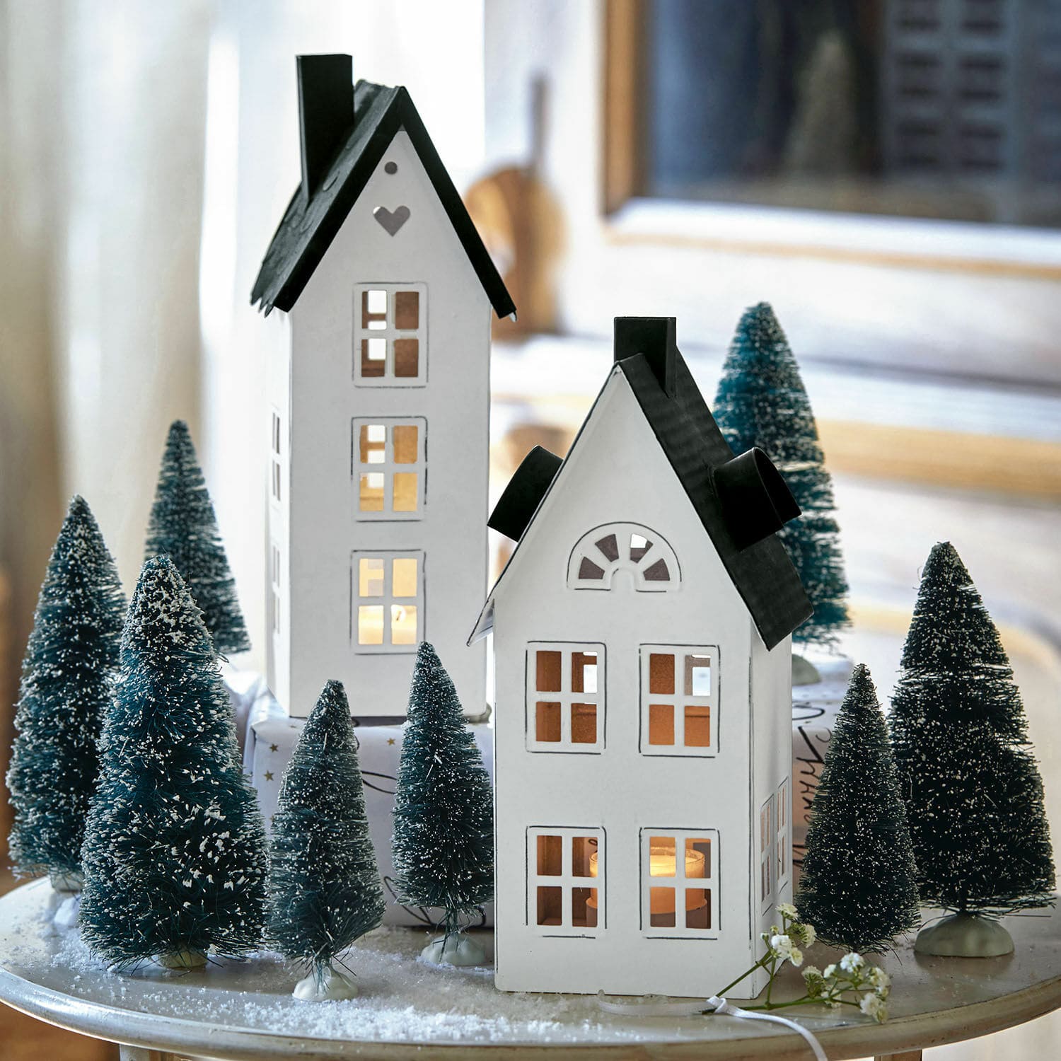 Lantaarn set van 2 Gunar Decoratieve witte metalen huisjes met zwarte daken omringd door kunstkerstbomen op een feestelijke tafel