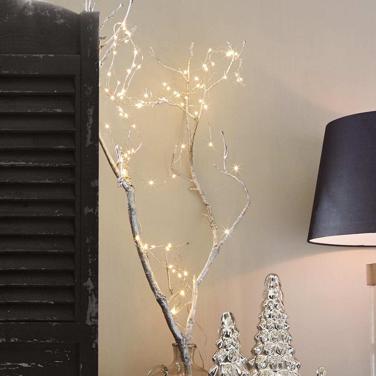 Decoratieve tak met LED-lichtjes in een glazen vaas naast zilveren kerstdecoraties en een zwarte lamp