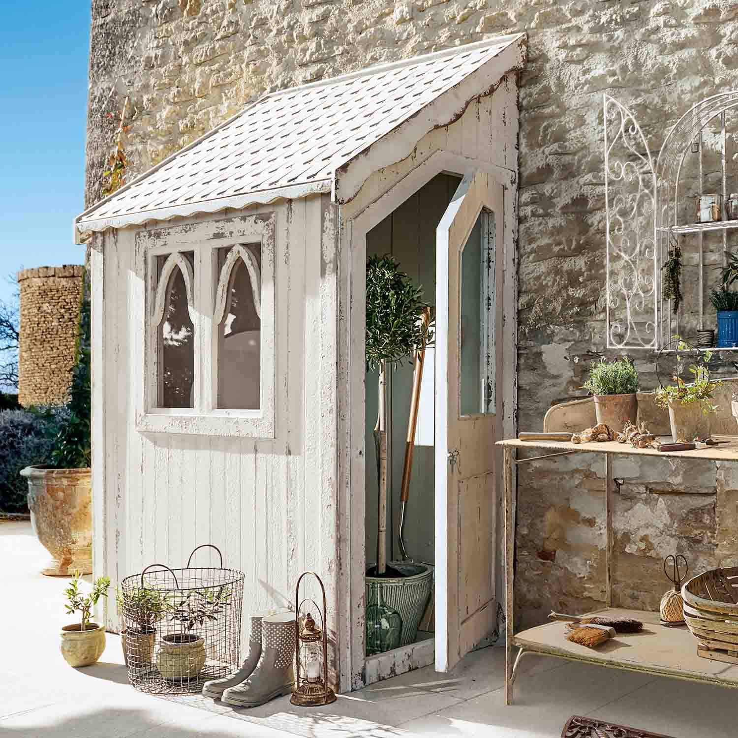Petit abri de jardin en bois blanc avec fenêtres et porte ouverte, outils et décorations de jardin