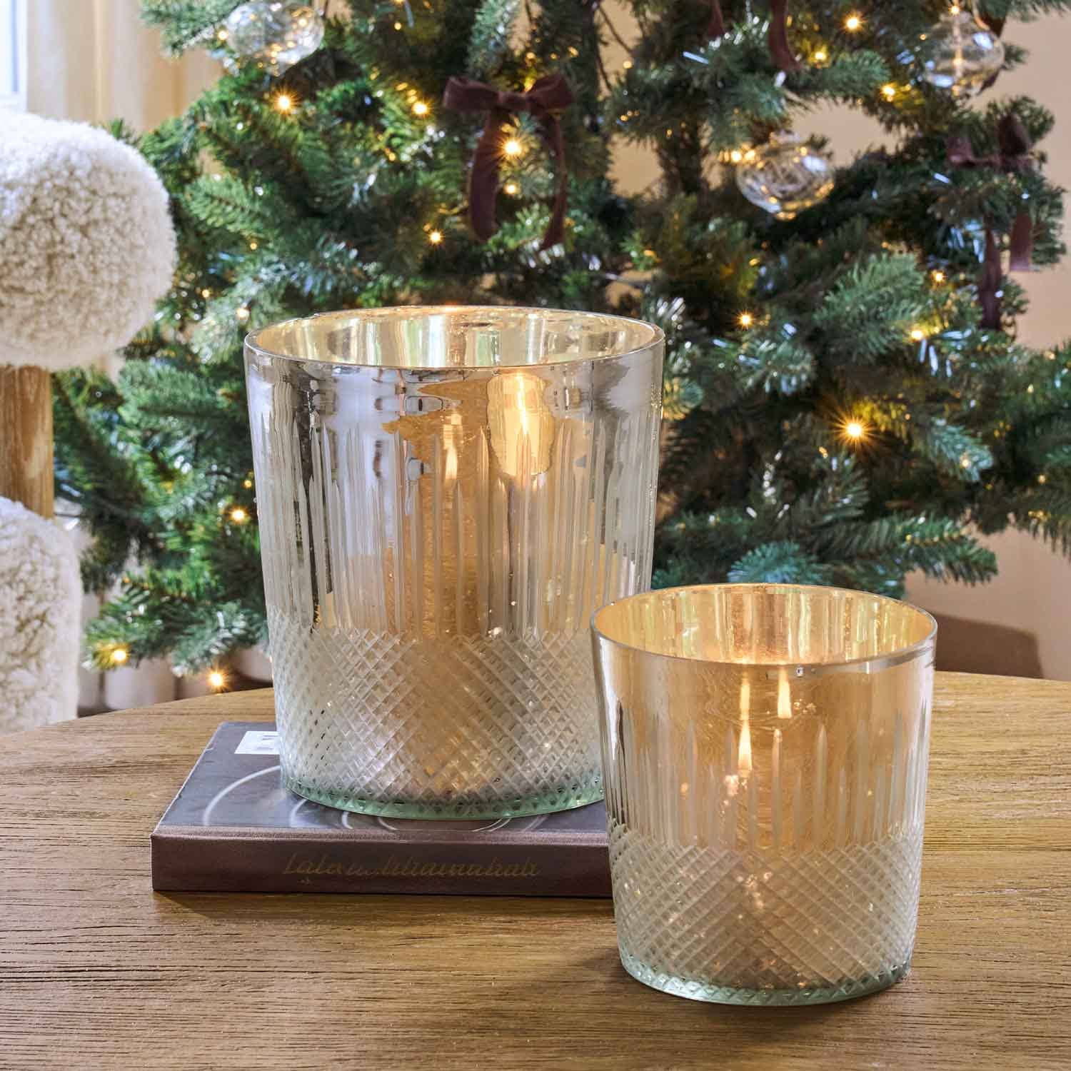 Deux photophores en verre à finition métallique sur une table en bois devant un sapin de Noël décoré