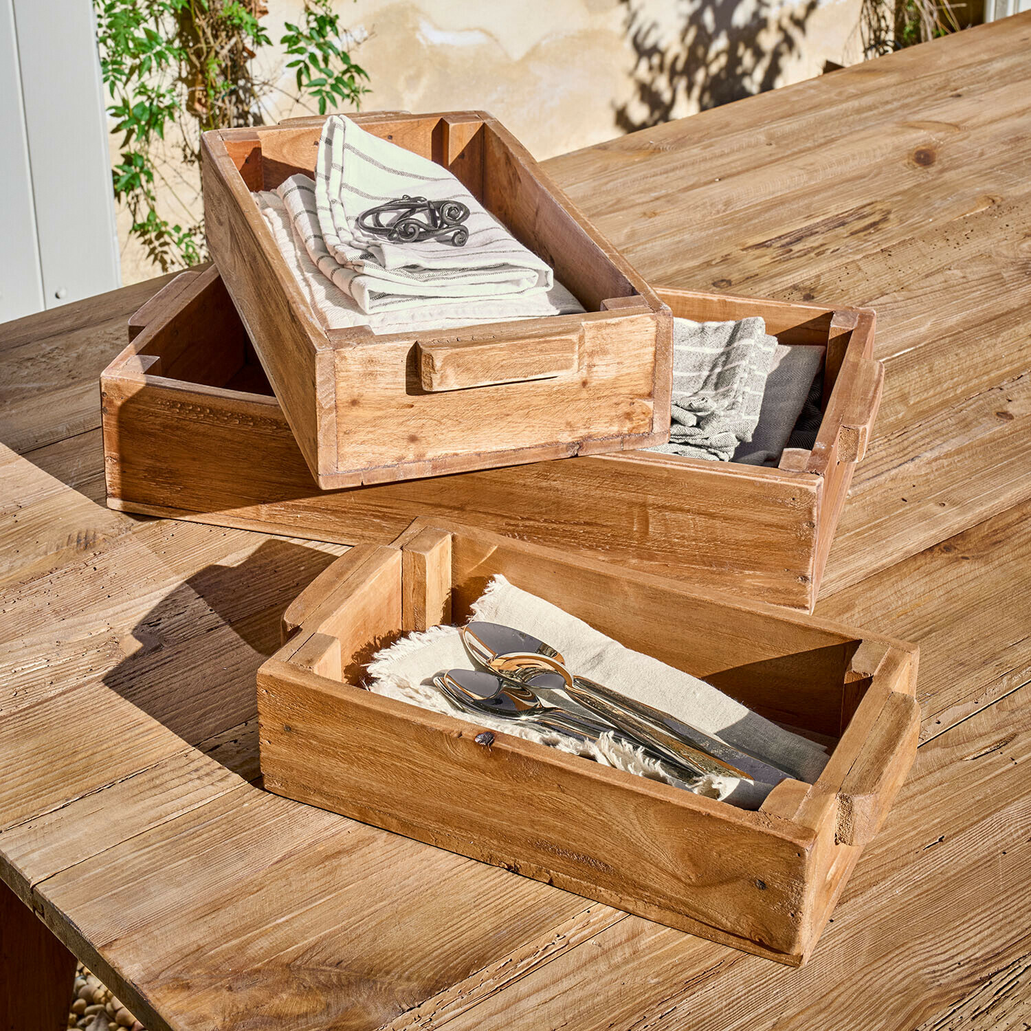 Lot de 3 boîtes Tarevo Ensemble de trois plateaux en bois naturel avec poignées, posés sur une table en bois, avec serviettes et couverts
