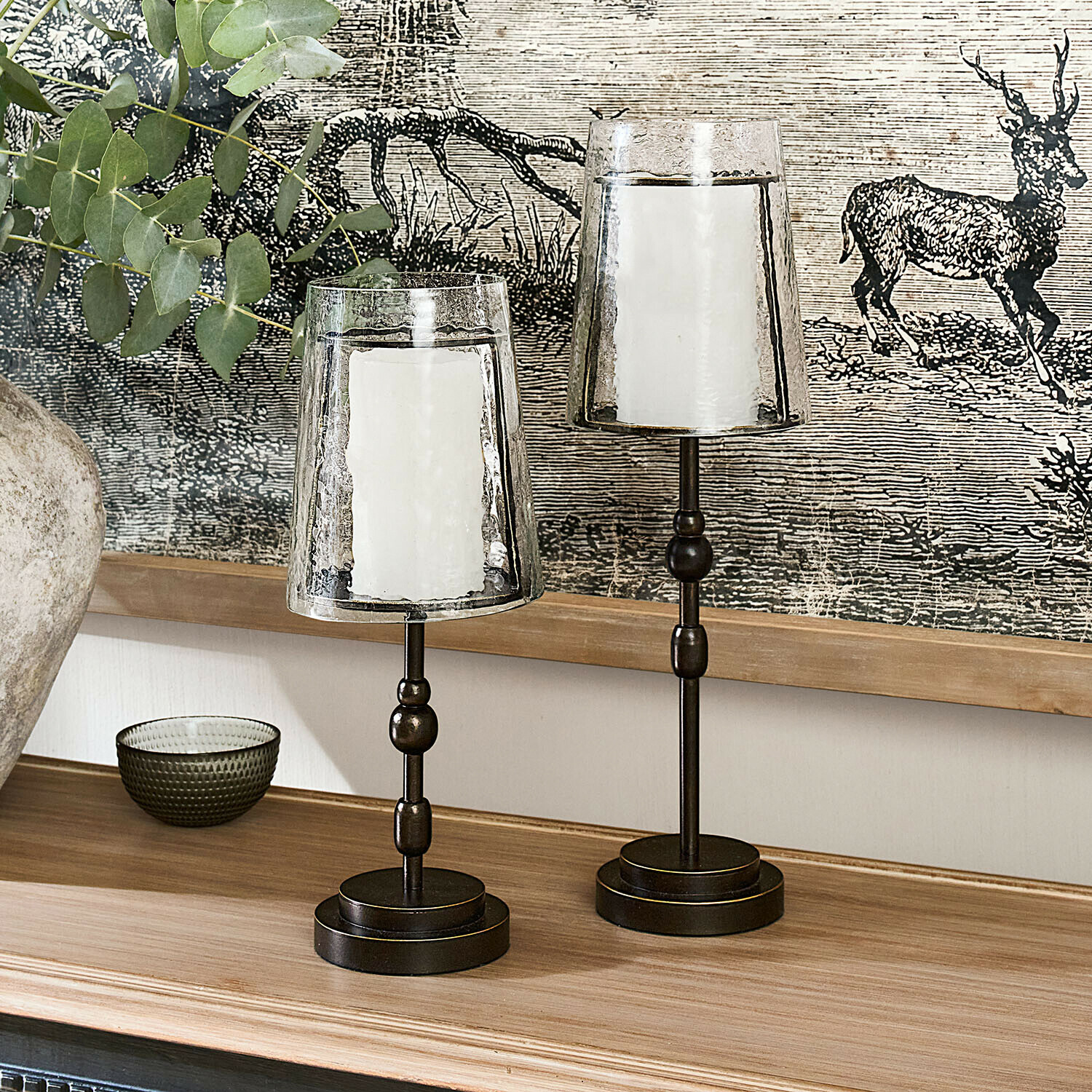 Twee metalen kandelaars met transparante glazen kappen en witte kaarsen op een houten blad