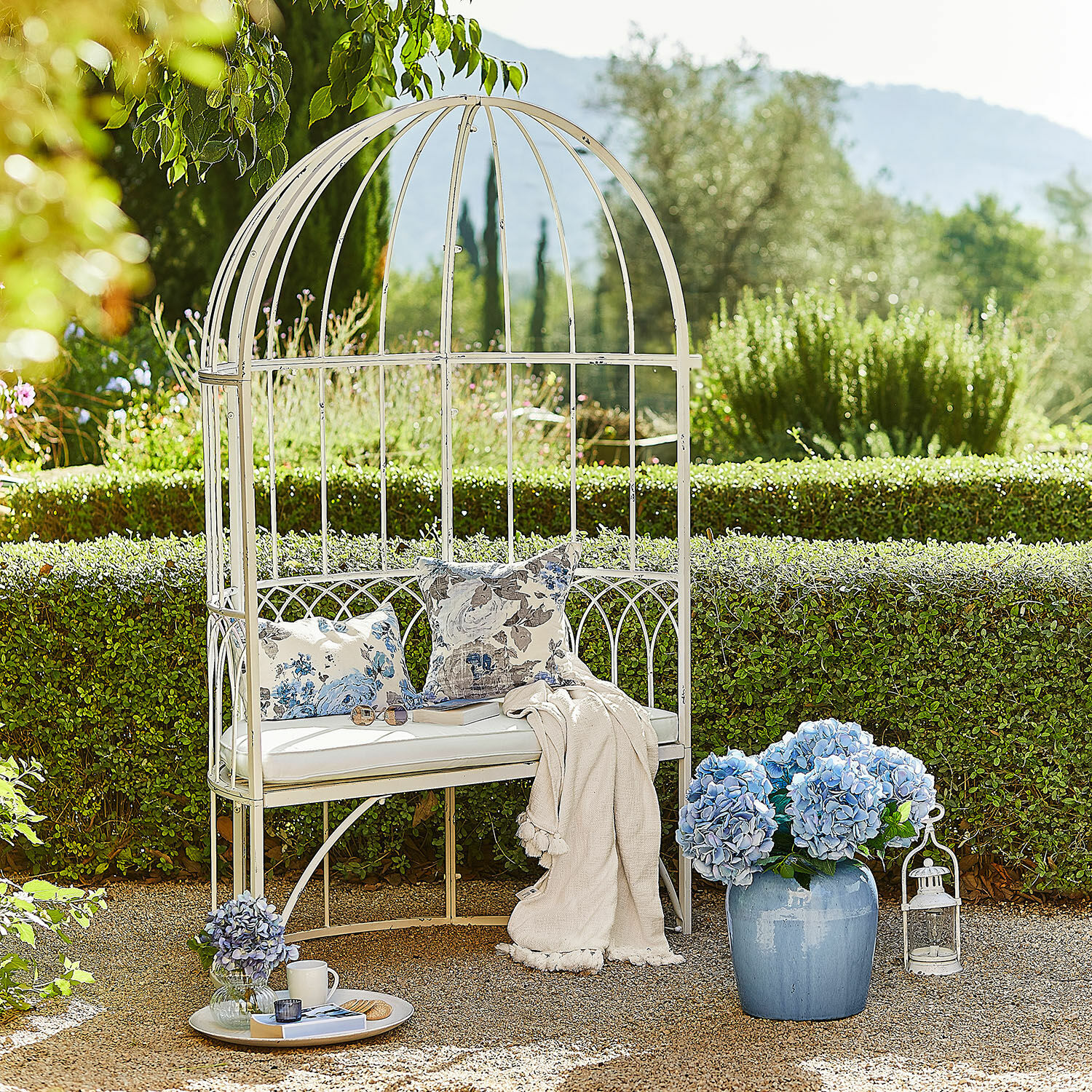 Bank Rivandelle Witte metalen tuinbank met kussens en deken, versierd met blauwe hortensia bloemen