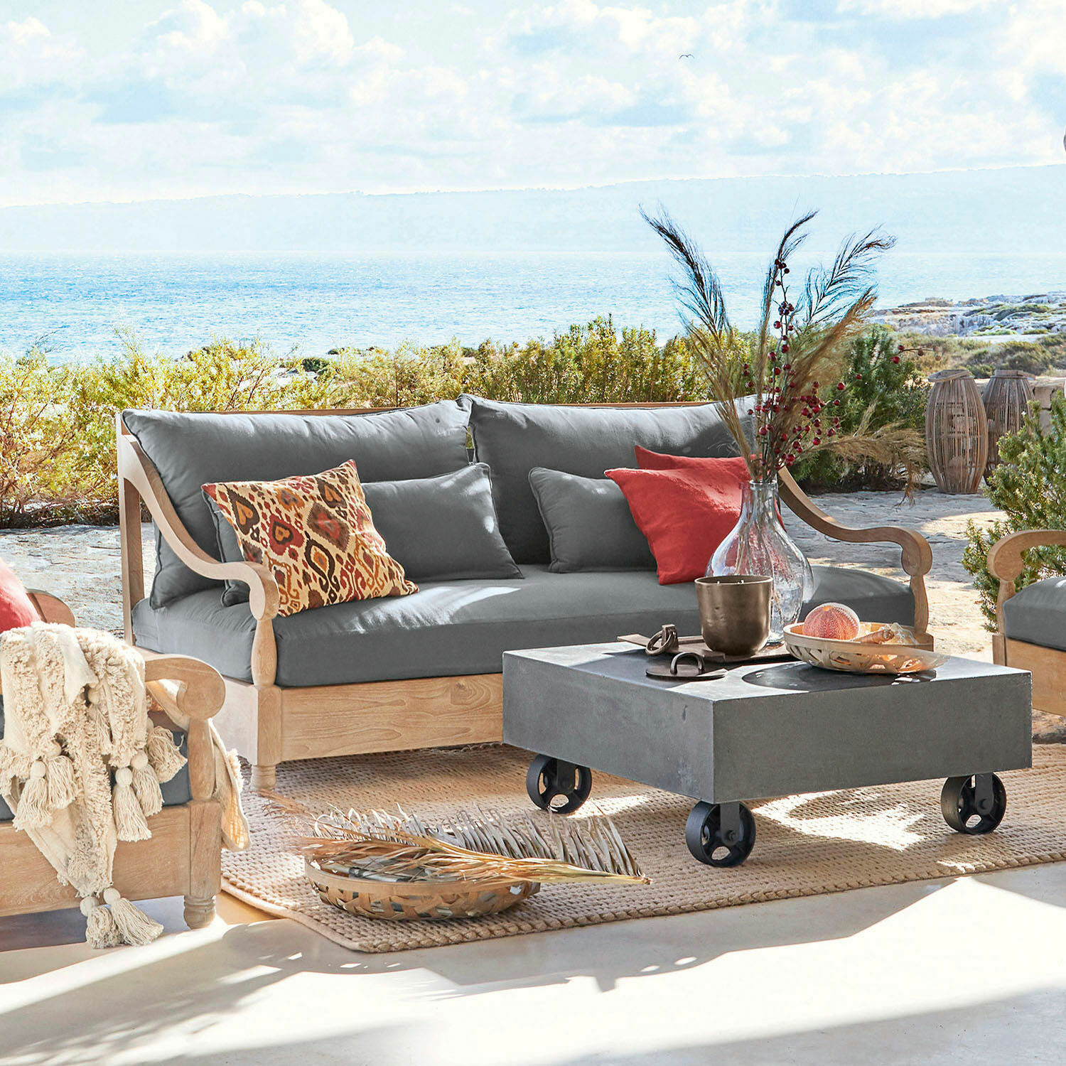 Canapé d'extérieur en bois clair avec coussins gris et coussins décoratifs colorés sur terrasse face à la mer