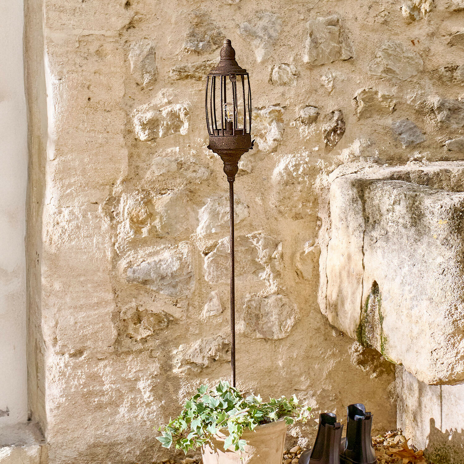 Lanterne LED rechargeable Lagia Lampe de jardin en métal rouillé sur pied long devant un mur en pierre rustique