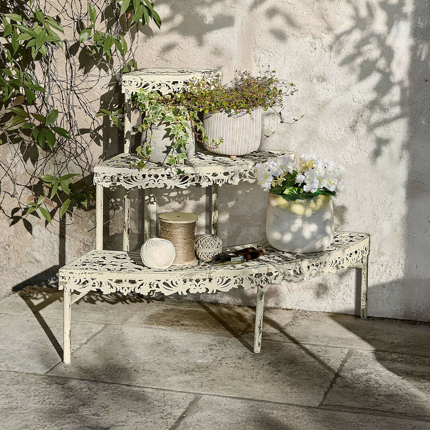 Plantentrapje Cresselay Crèmekleurige metalen etagère met opengewerkt patroon, planten en tuingereedschap op stenen terras