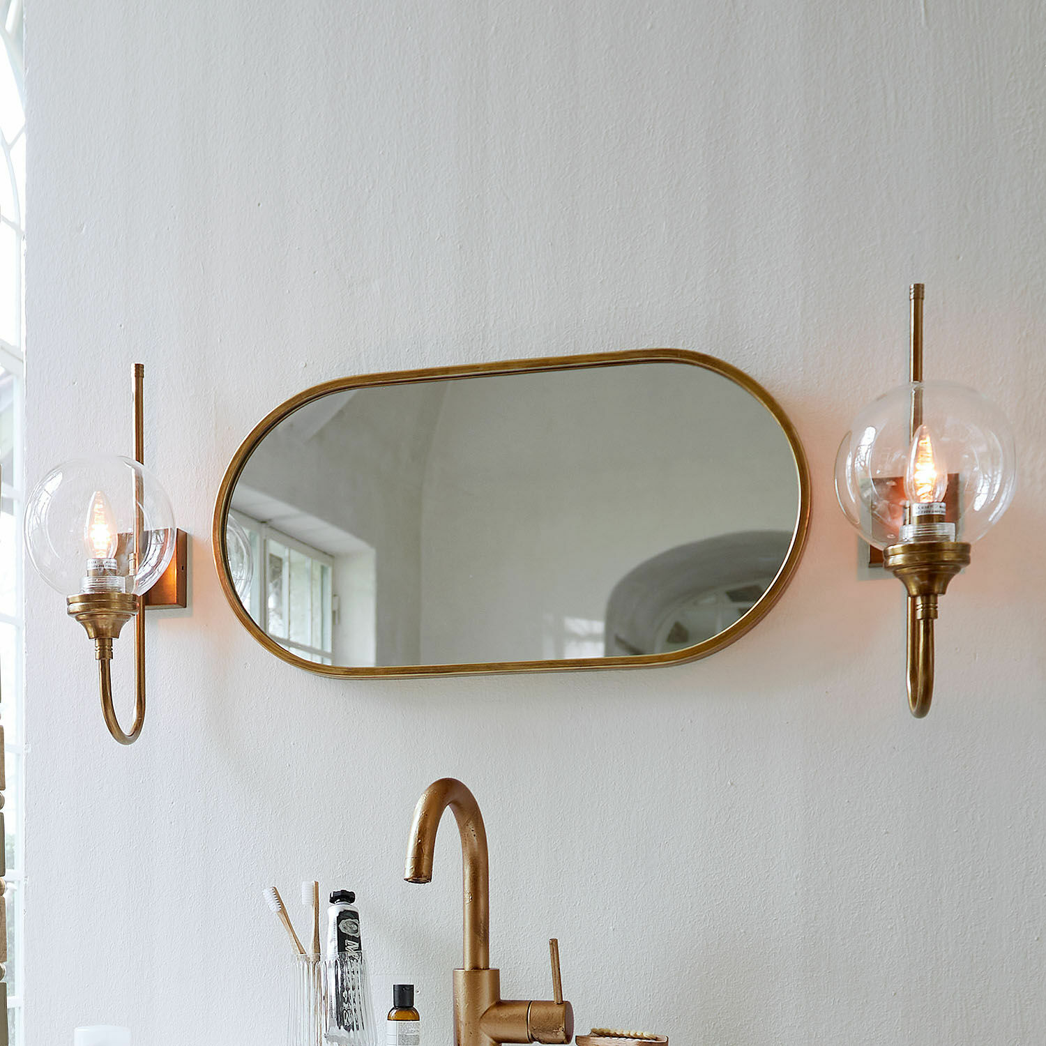 Miroir Anthé Miroir ovale avec cadre en laiton et deux appliques murales à abat-jour transparent dans une salle de bain