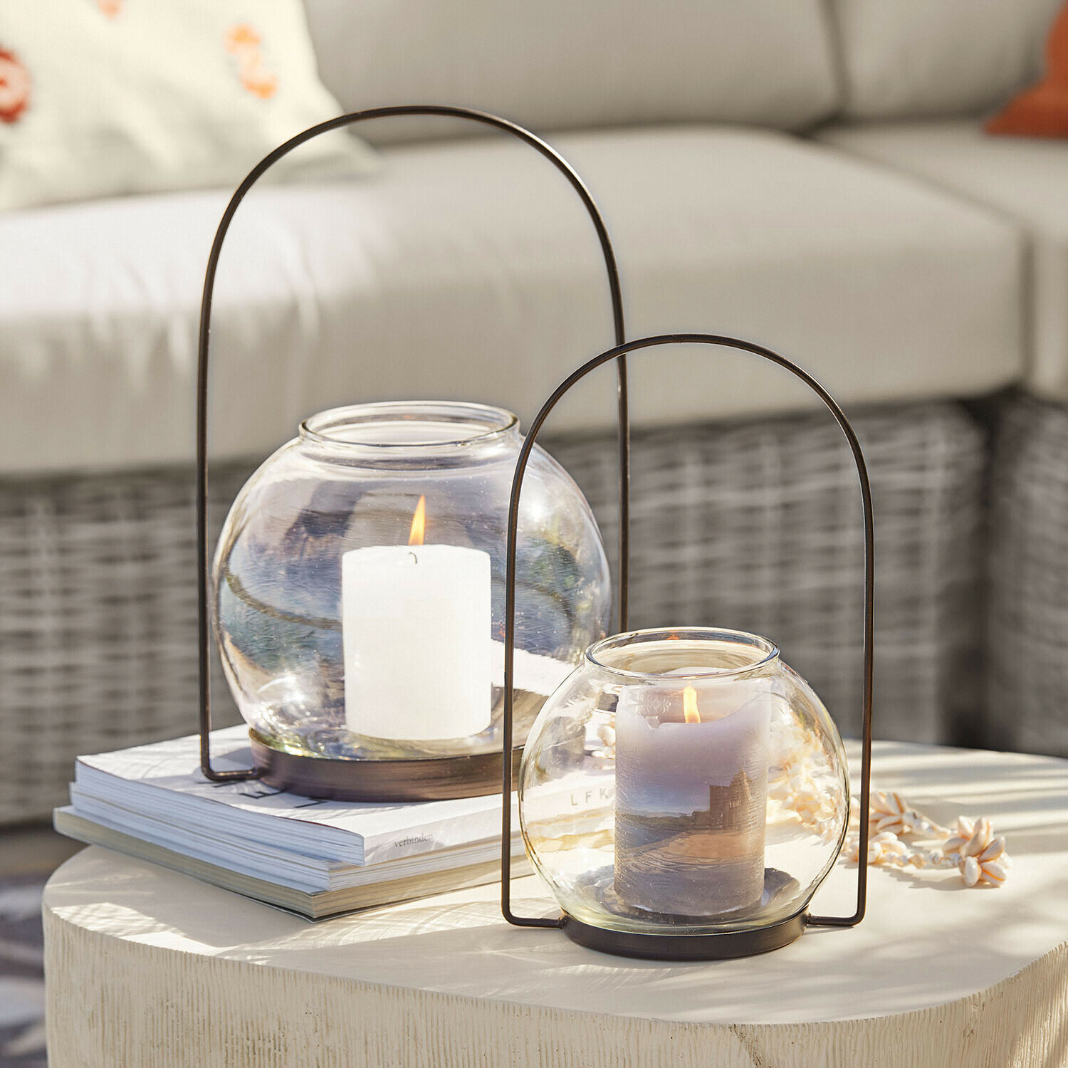 Lot de 2 photophores Serenaro Deux lanternes en verre avec poignées métalliques et bougies allumées sur une table claire décorative