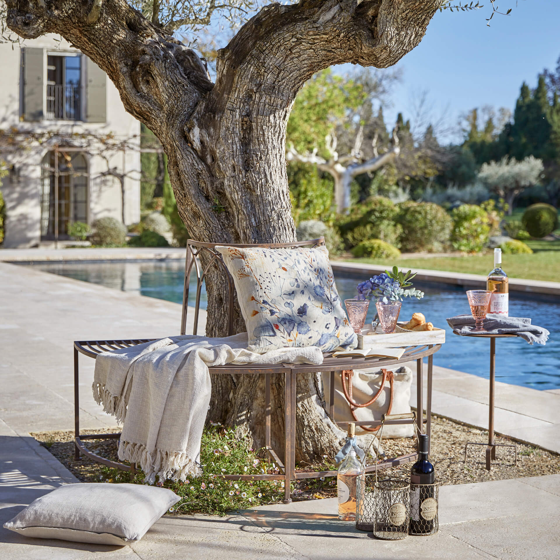Banc en métal couleur cuivre sous un arbre avec coussin et plaid près de la piscine dans un jardin