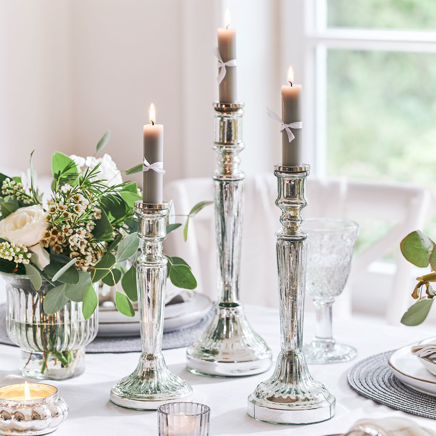 Trois chandeliers argentés avec bougies grises sur une table blanche décorée de fleurs et de verre