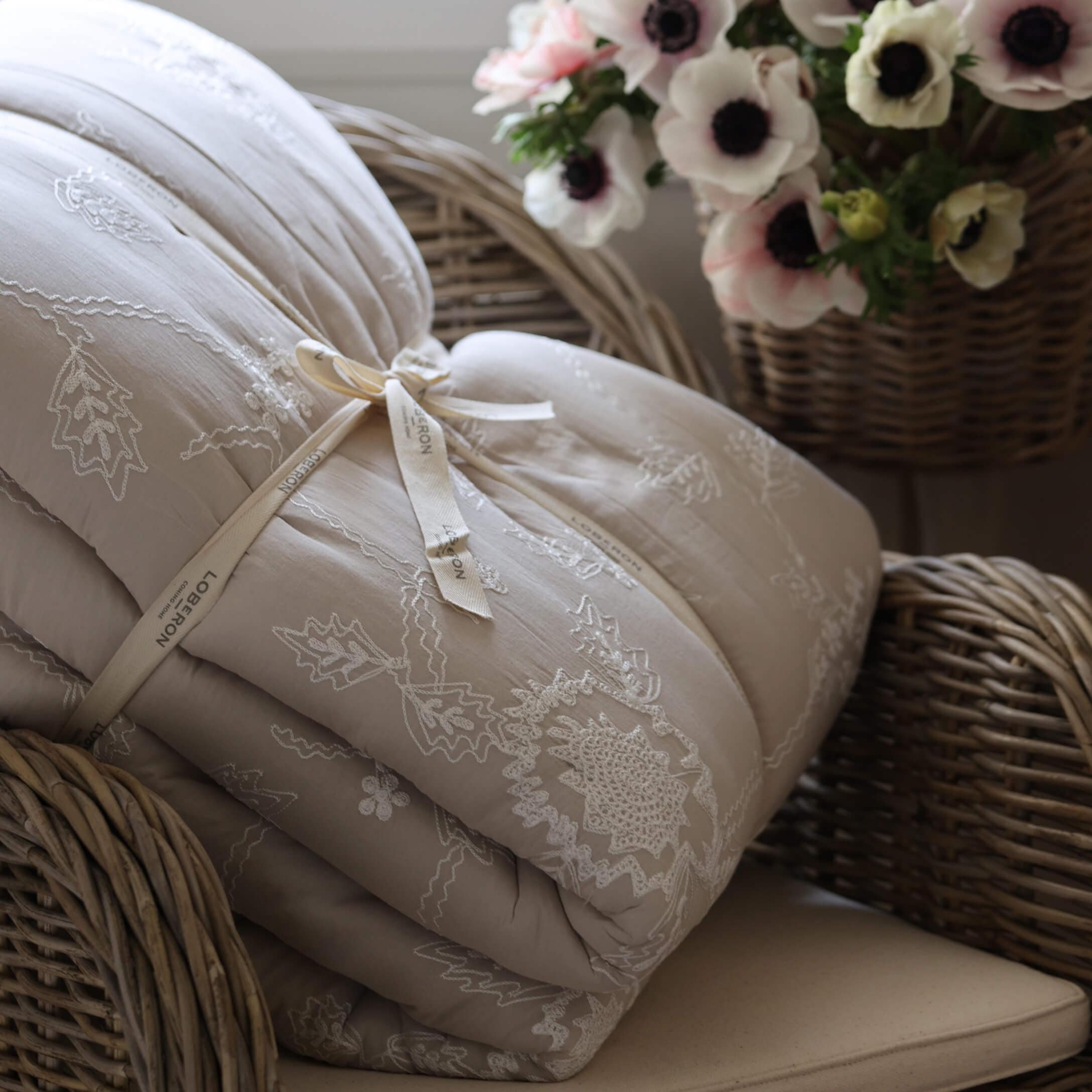 Beige gewatteerde deken met borduurwerk op een rotan stoel naast een bloemenmand, sfeervolle woondecoratie