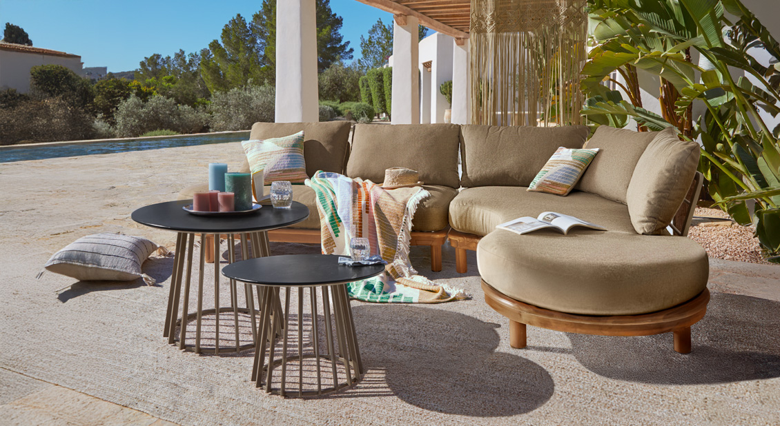 Canapé d'angle beige avec base en bois en extérieur, deux tables basses noires et coussins colorés