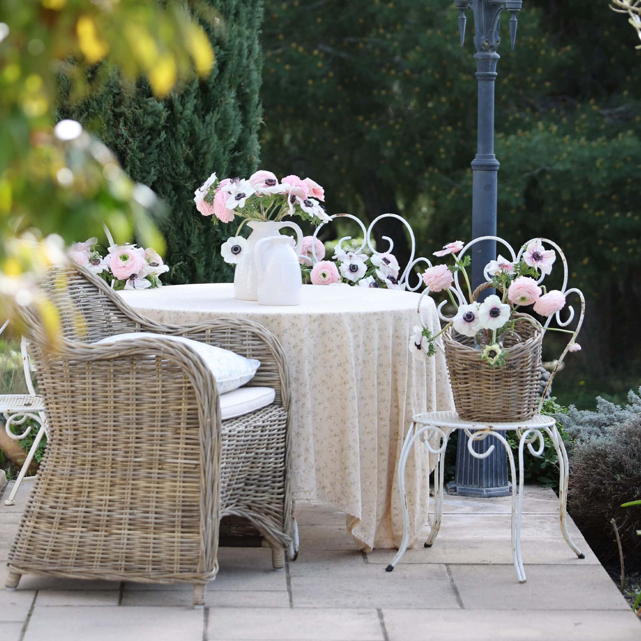 Coin de jardin avec fauteuil en osier, table blanche avec nappe et fleurs dans des vases, ambiance romantique