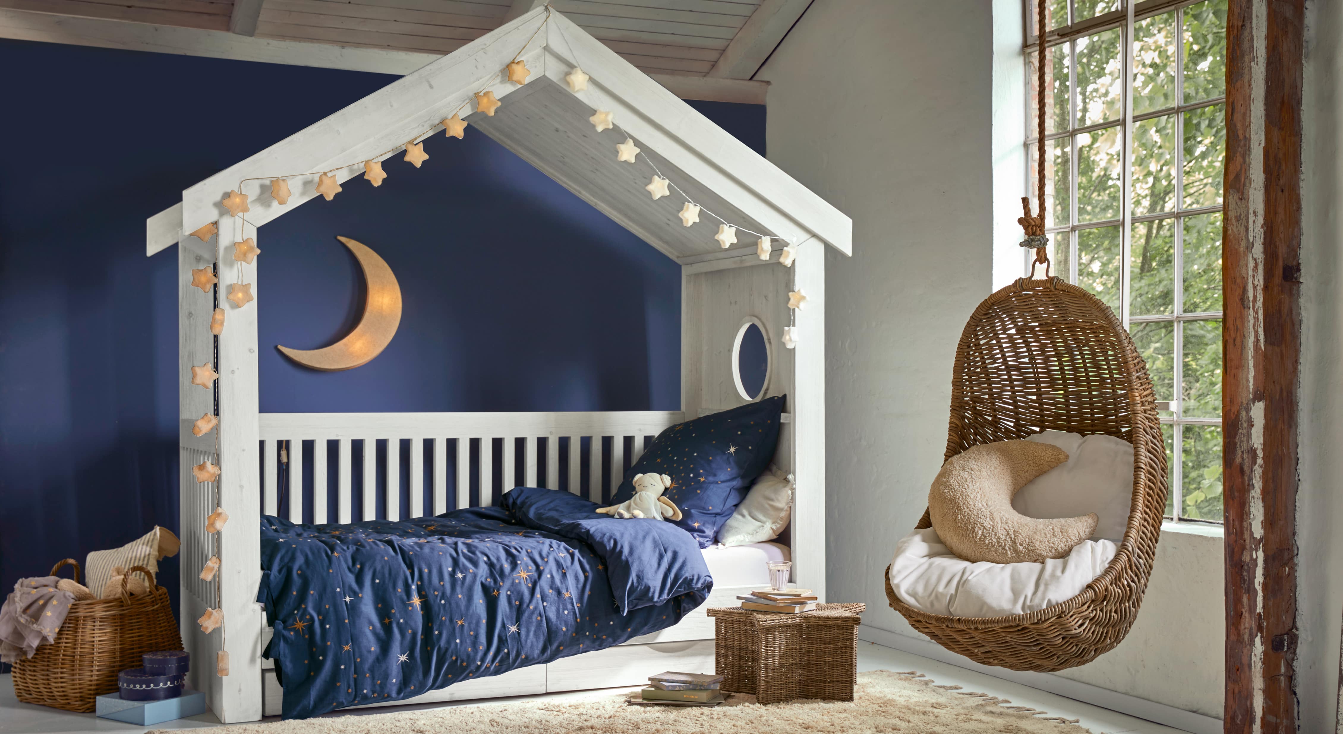 Lit en bois blanc en forme de maison avec literie bleue étoilée et fauteuil suspendu en osier dans une chambre d'enfant