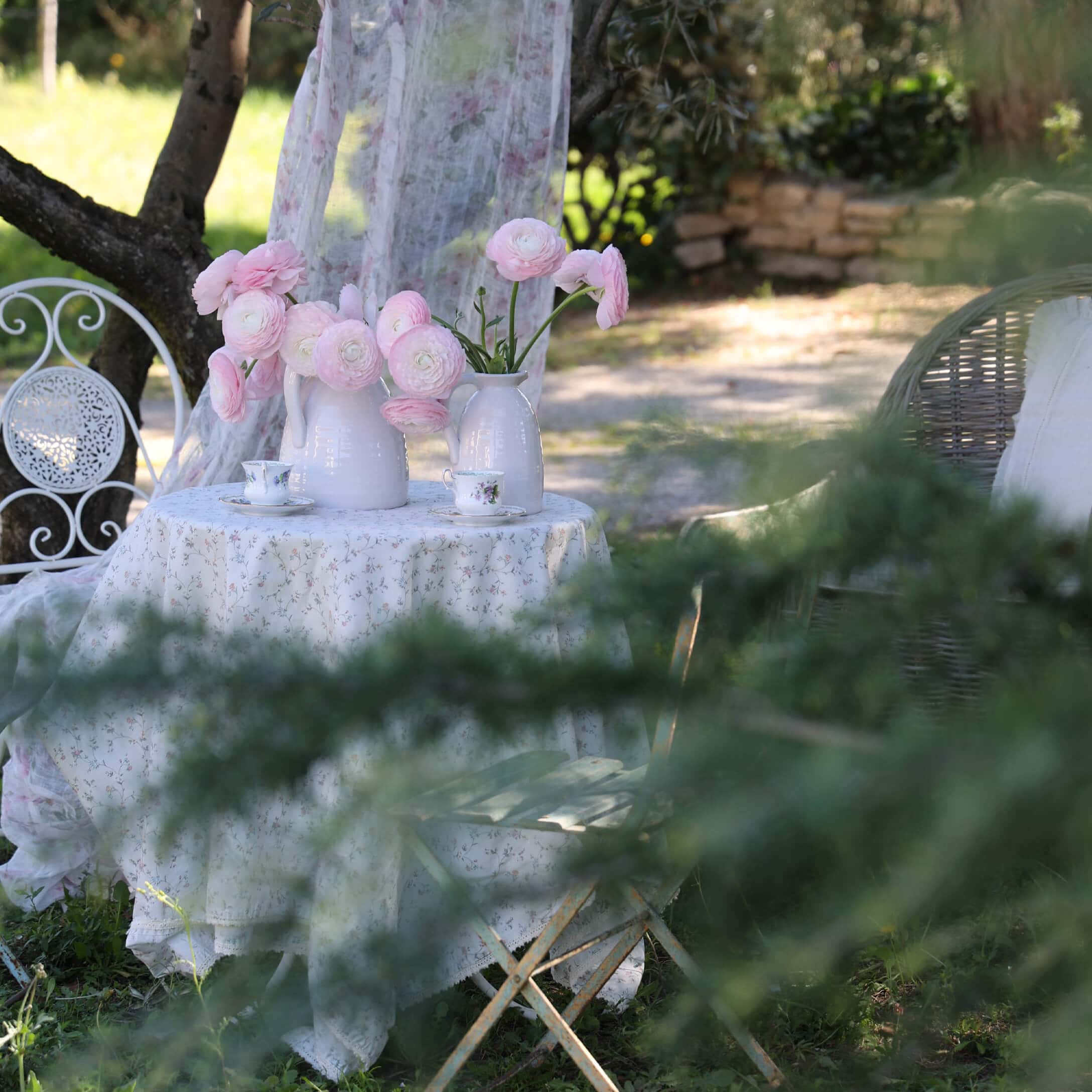 Table de jardin blanche avec nappe fleurie et vases blancs contenant des fleurs roses, ambiance romantique