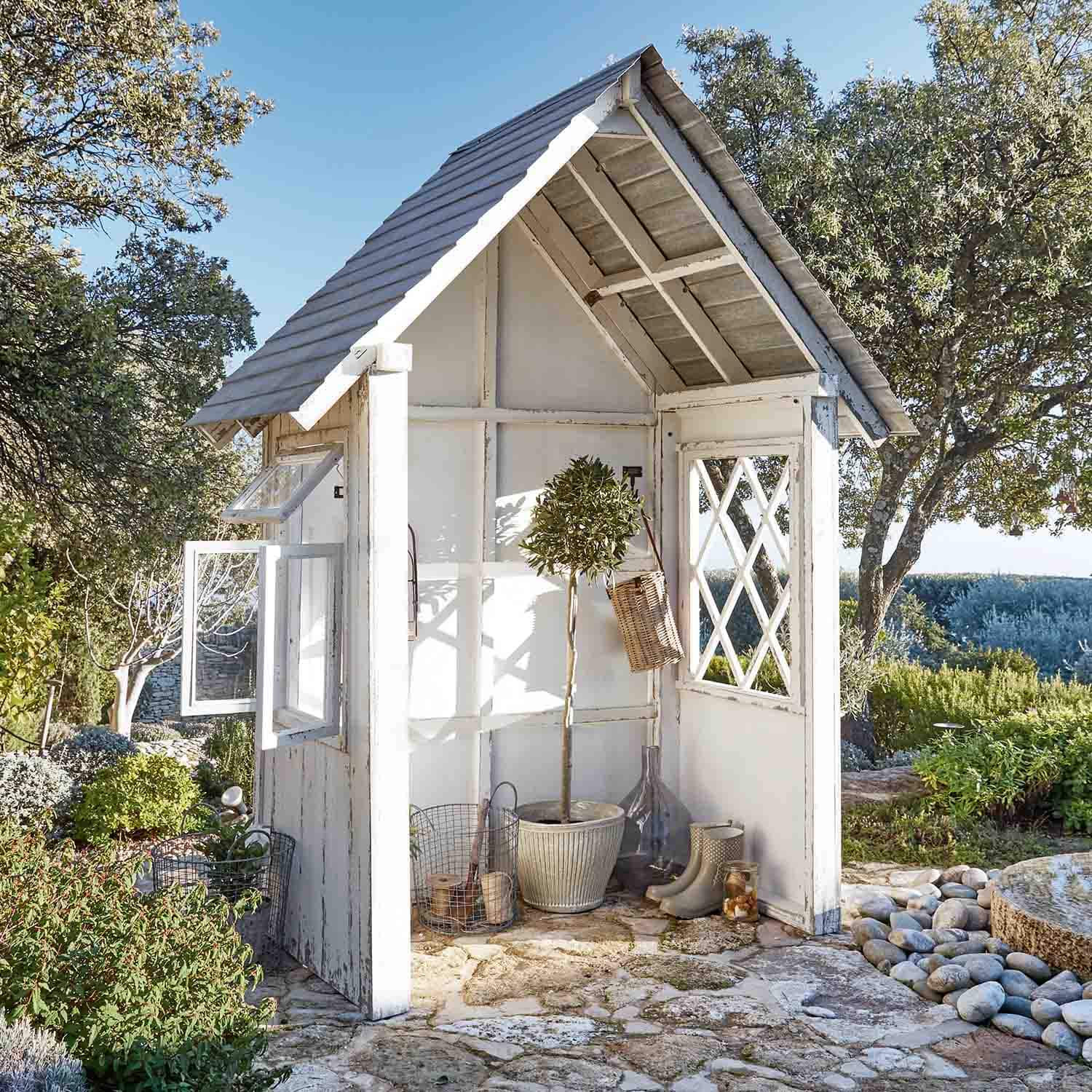 Petit abri de jardin en bois blanc avec fenêtres et plantes sur sol en pierre dans un jardin ensoleillé