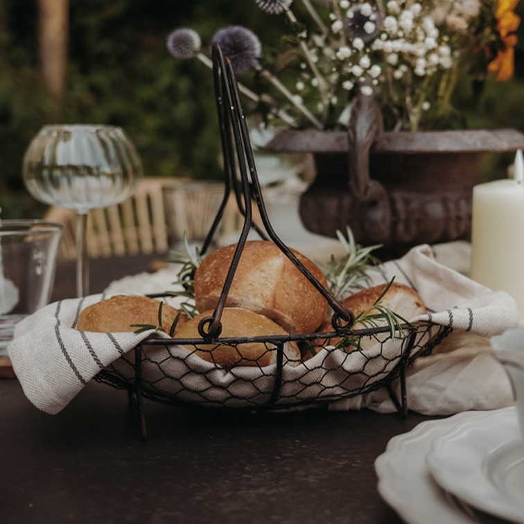 Panier en métal avec poignées, doublé d’un torchon en lin, contenant des petits pains et des herbes sur une table