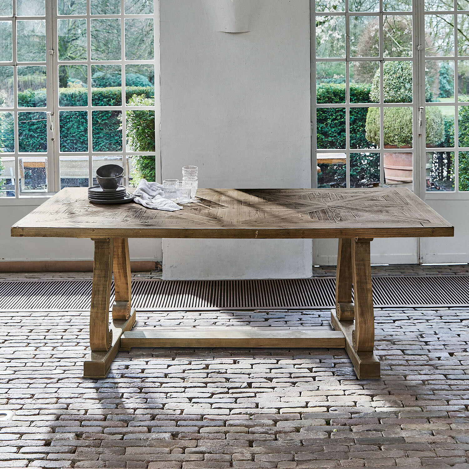 Table Campbelltown Table en bois naturel avec plateau décoratif devant de grandes fenêtres et un sol en briques