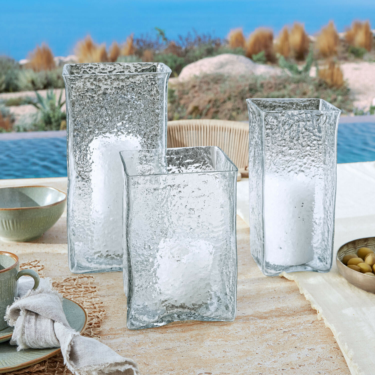Lot de 3 photophores Xarlac Trois lanternes en verre transparent texturé avec bougies blanches posées à l'extérieur près de la piscine