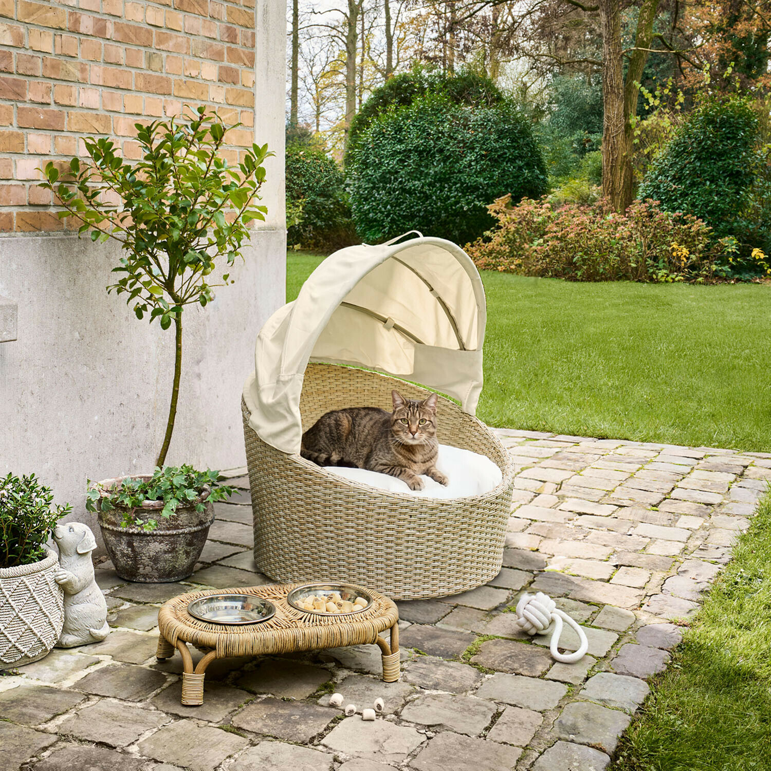 Ovaal kattenmandje van beige rattan met wit kussen en kap, geplaatst op een stenen terras buiten