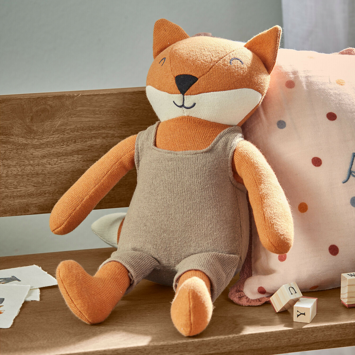 Zachte stoffen vos in beige outfit zittend op houten bank met houten leerblokken