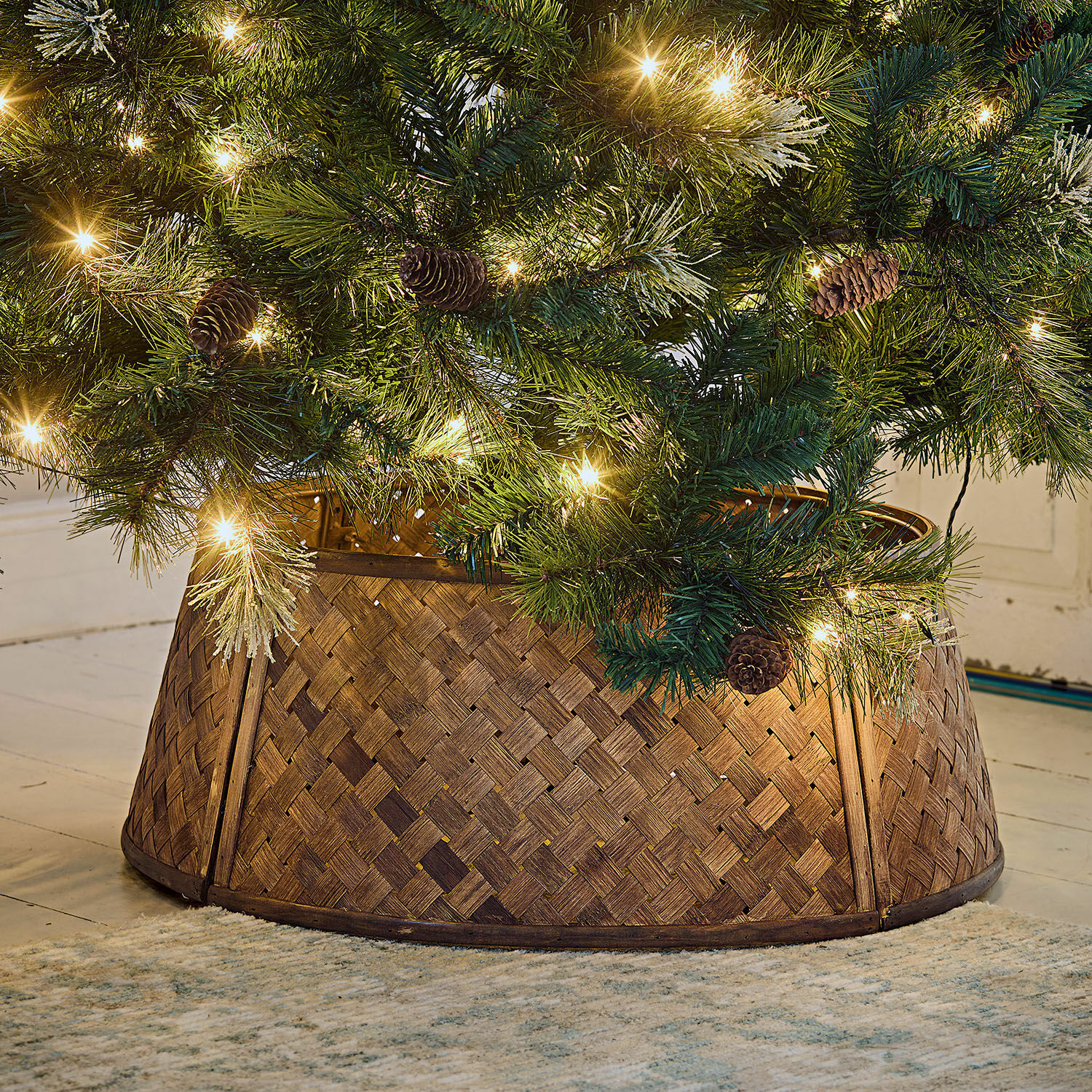 Cache-pied pour sapin de Noël Eden Support de sapin en bois tressé marron sous un sapin vert décoré de lumières et pommes de pin