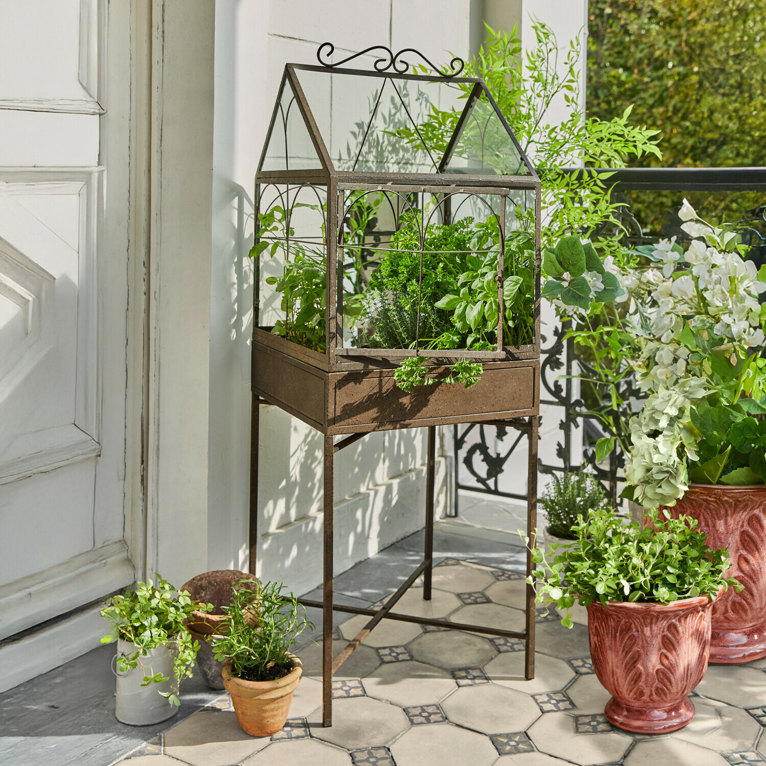 Metalen kas met roestkleurige afwerking op standaard met groene kruiden op betegeld terras