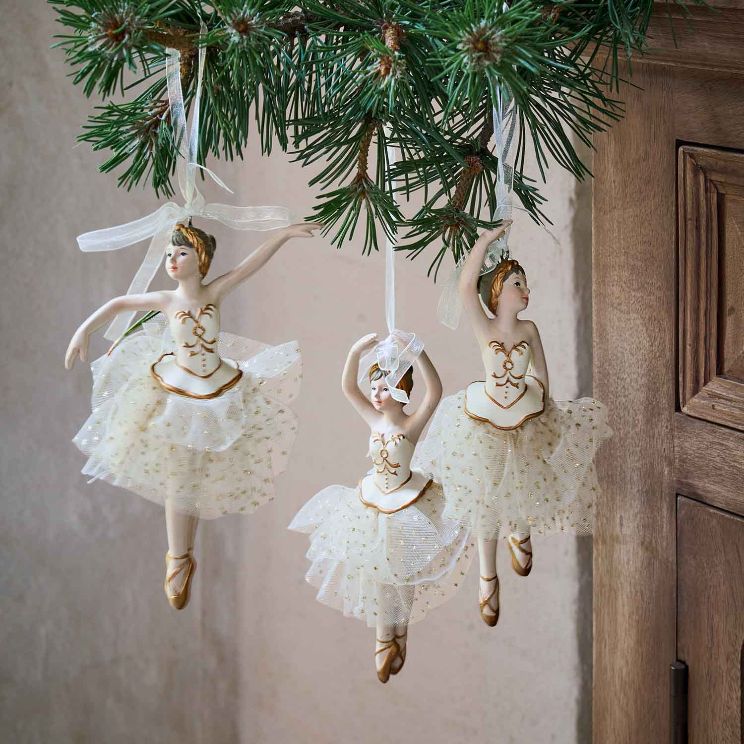 Drie witte ballerina-ornamenten met tule rokken en gouden details hangend aan een dennentak