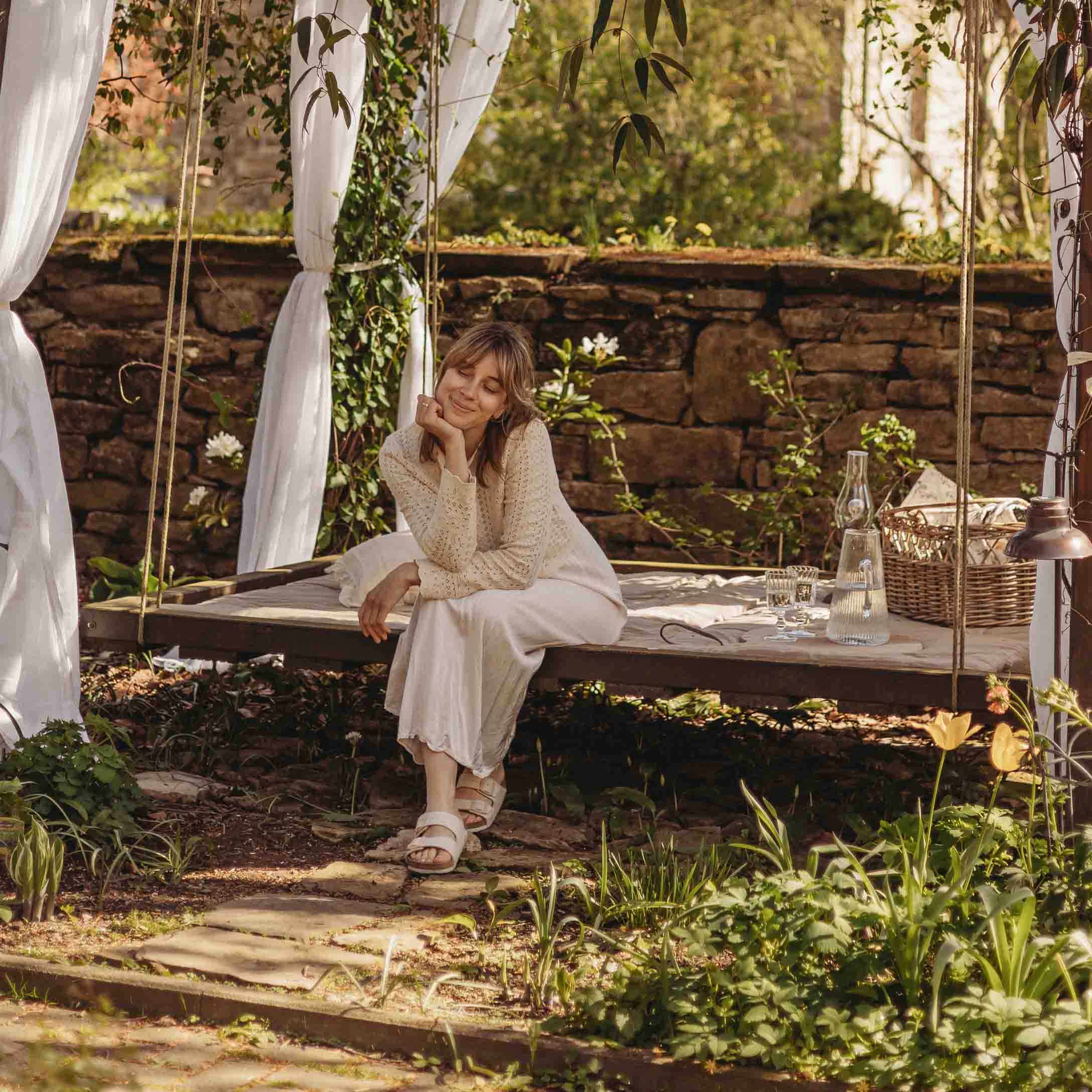 Femme en pull beige assise sur une balançoire de jardin en bois avec rideaux blancs et décor naturel