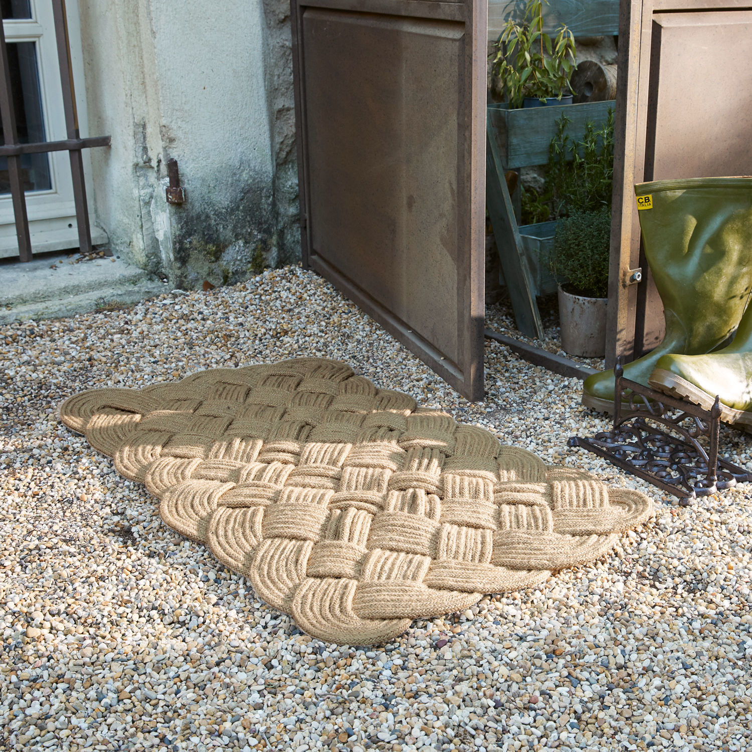 Beige deurmat van gevlochten touw op grind bij open deur met groene laarzen ernaast