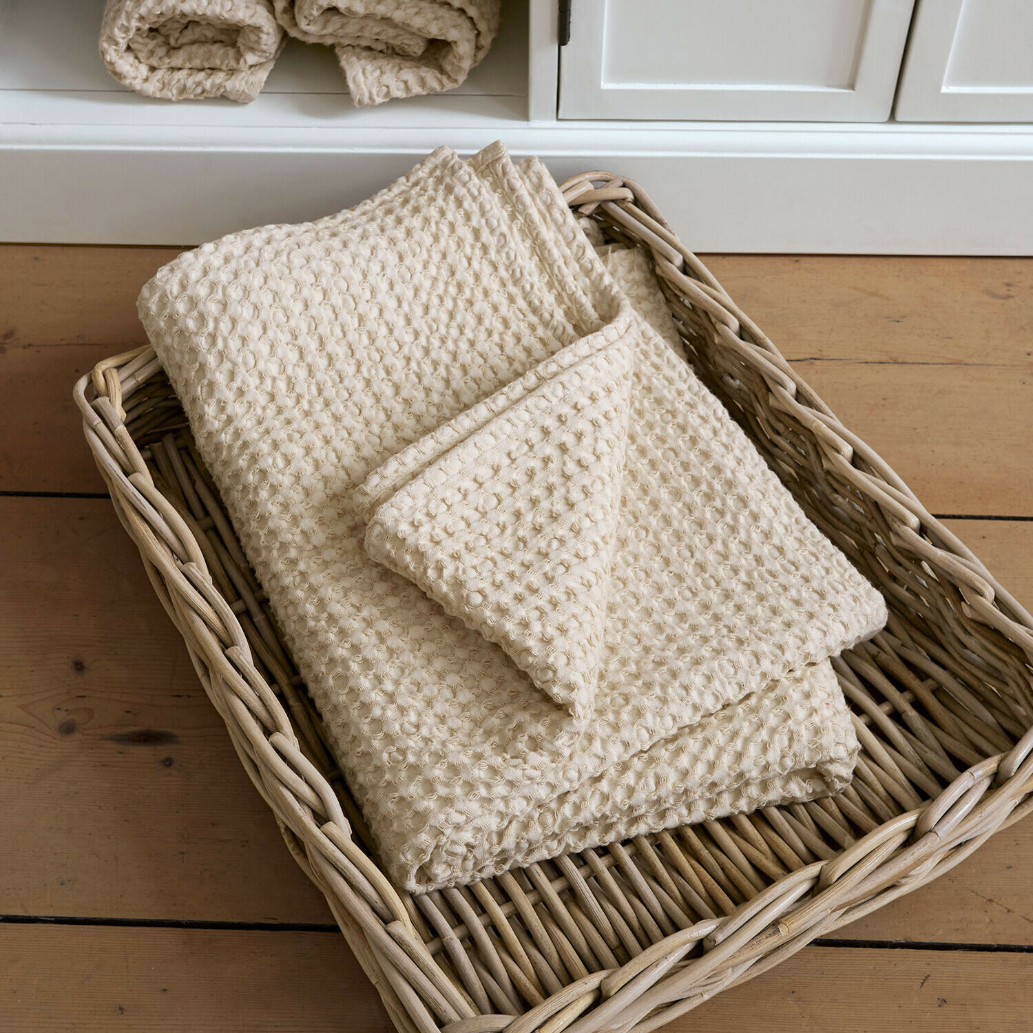 Serviette de bain Maralei Plaid crème en coton gaufré dans un panier en osier posé sur un sol en bois clair