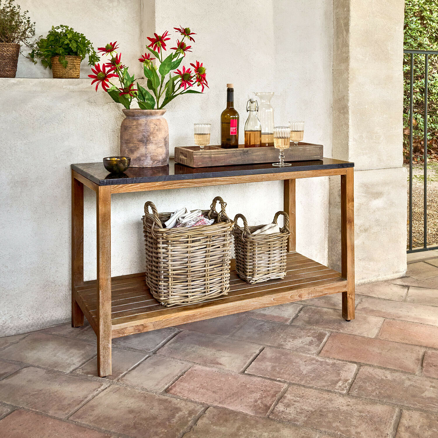 Table de cuisine d’extérieur Corviano Console en bois avec plateau noir, décorée d'un vase à fleurs rouges et de paniers en osier sous l'étagère