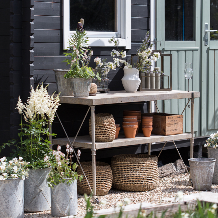 Metalen tuintafel met bloempotten, rieten manden en bloemen voor een zwarte muur en groene deur