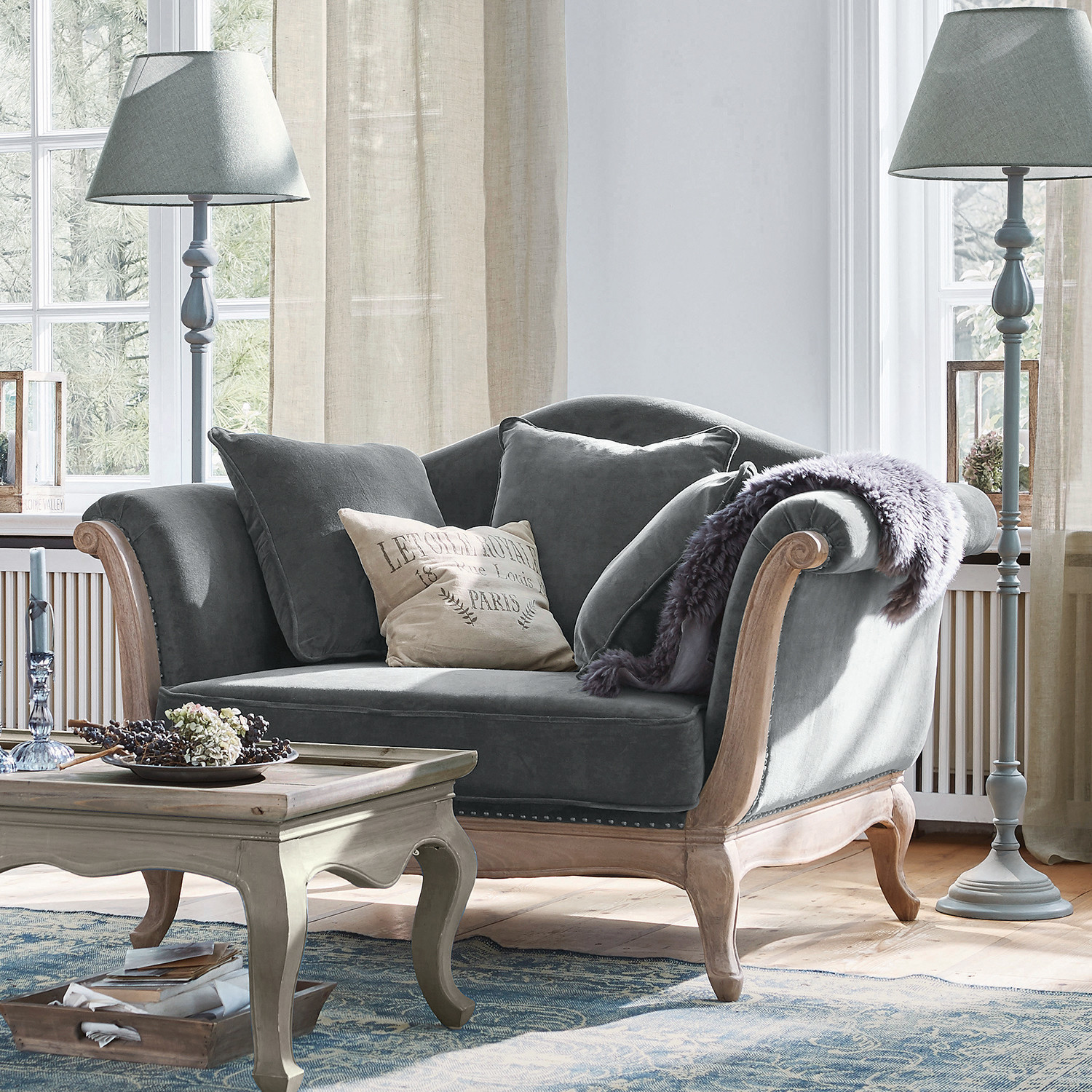 Fauteuil Sumerset Canapé gris en velours avec cadre en bois et coussins dans un salon lumineux avec lampes sur pied