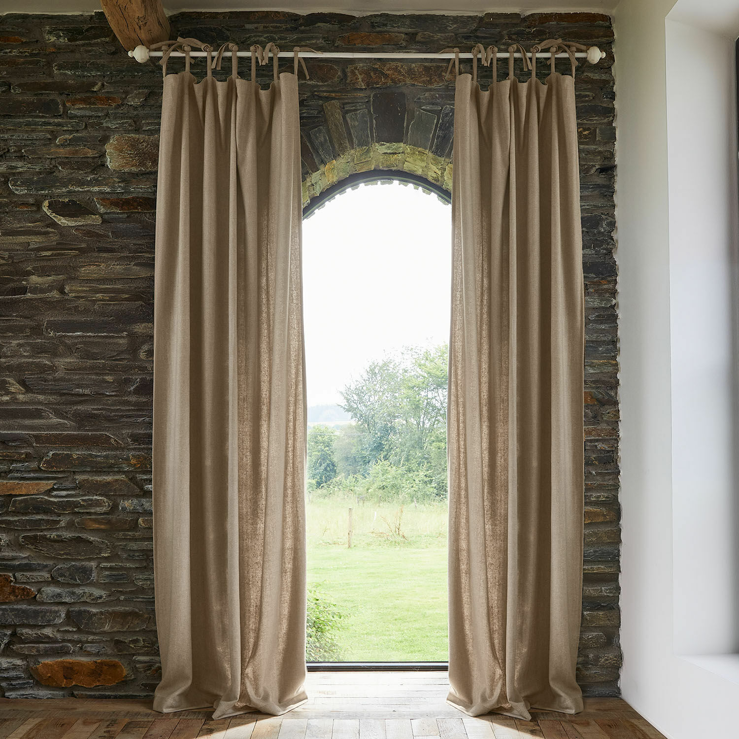 Gordijn Seleneia Beige gordijnen van natuurlijke stof aan roede in stenen interieur met uitzicht op tuin