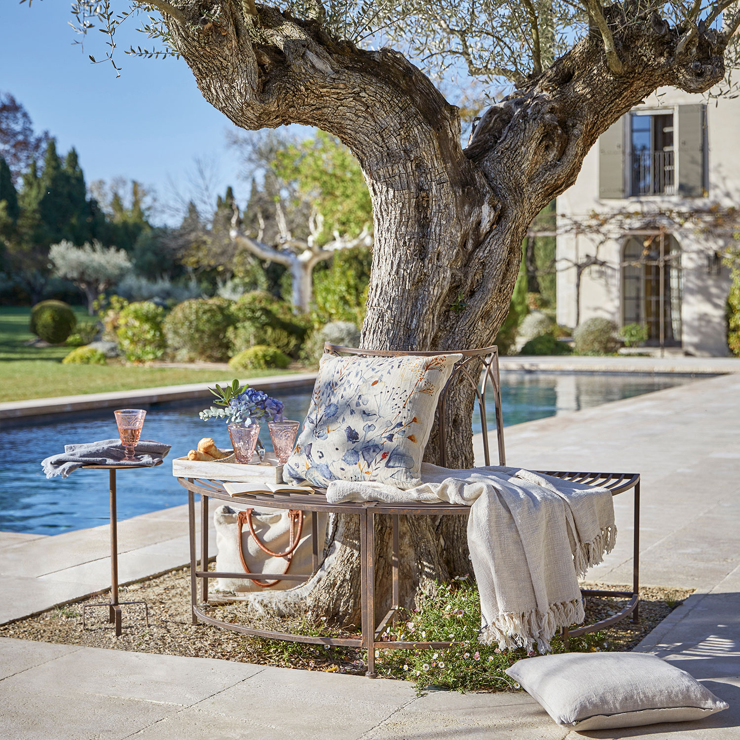 Banc en métal autour d'un arbre avec coussin et plaid près d'une piscine dans un jardin