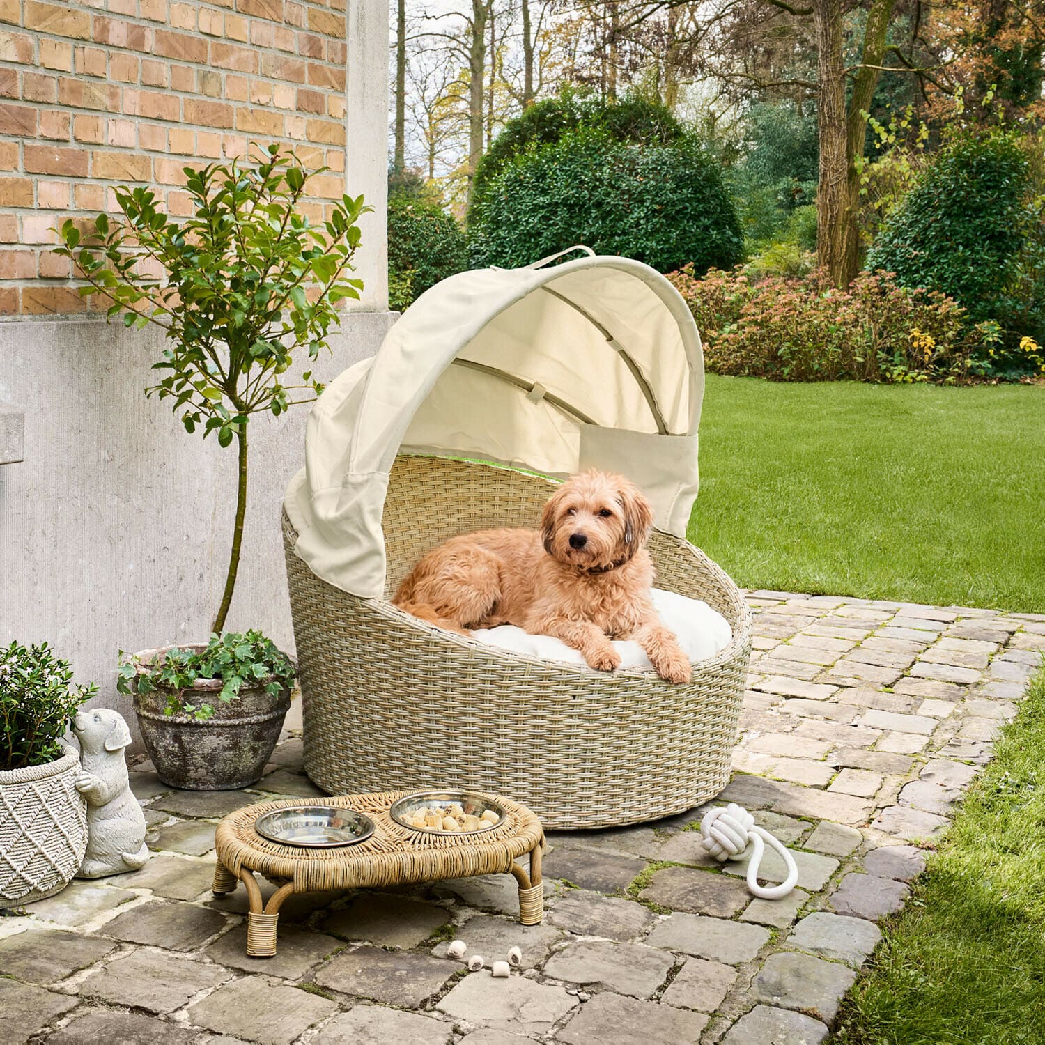 Panier rond en rotin beige avec auvent crème et coussin pour chien dans un jardin pavé