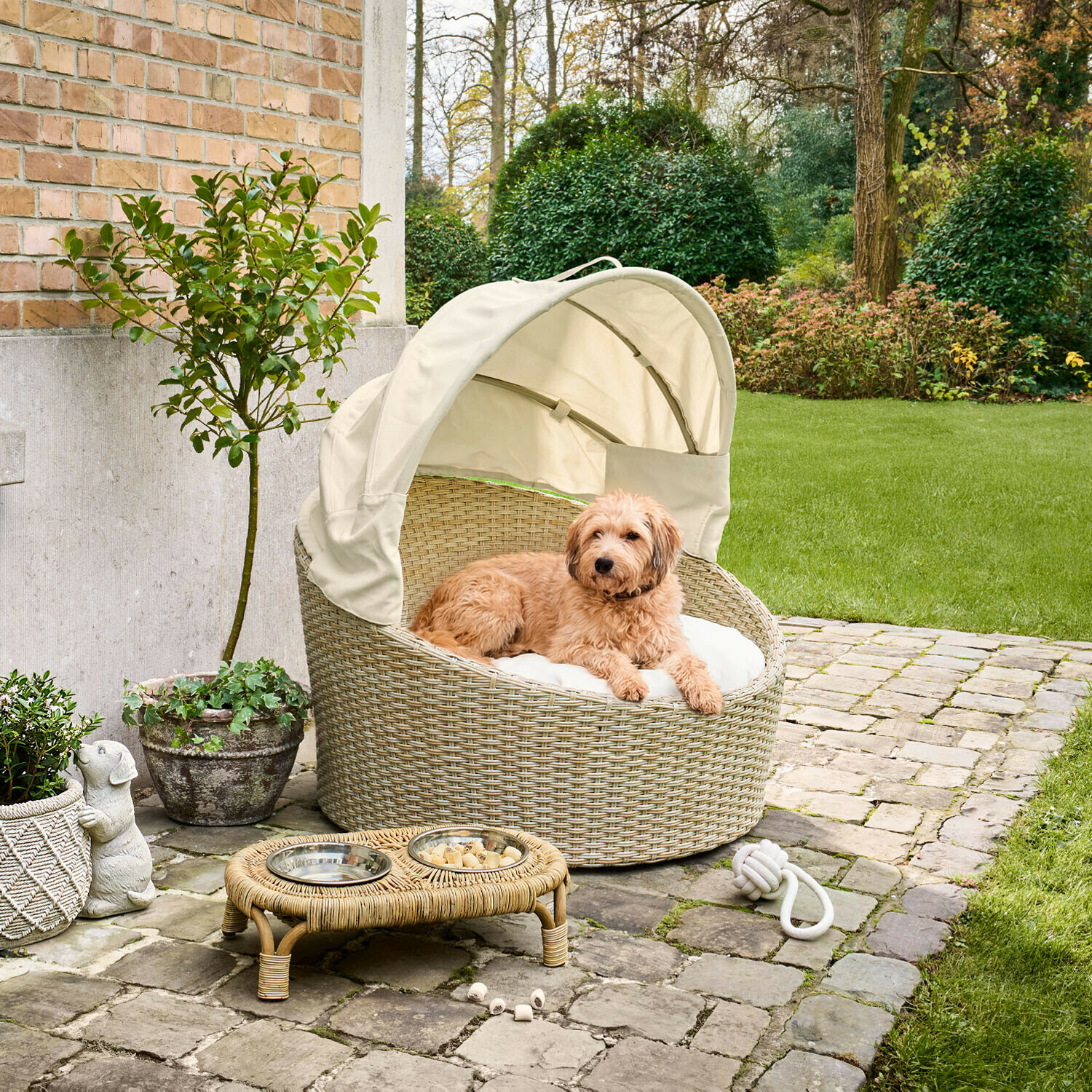 Mand voor huisdieren voor binnen en buiten Brinelza Ronde hondenmand van beige rattan met crèmekleurige kap en kussen op een betegeld terras
