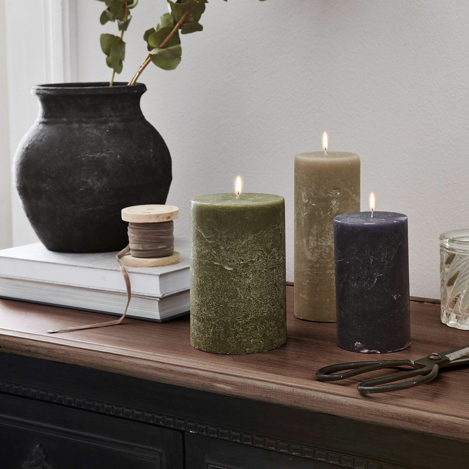 Lot de 3 bougies Pretin Trois bougies allumées vert, beige et noir sur un plateau en bois avec décorations d'intérieur